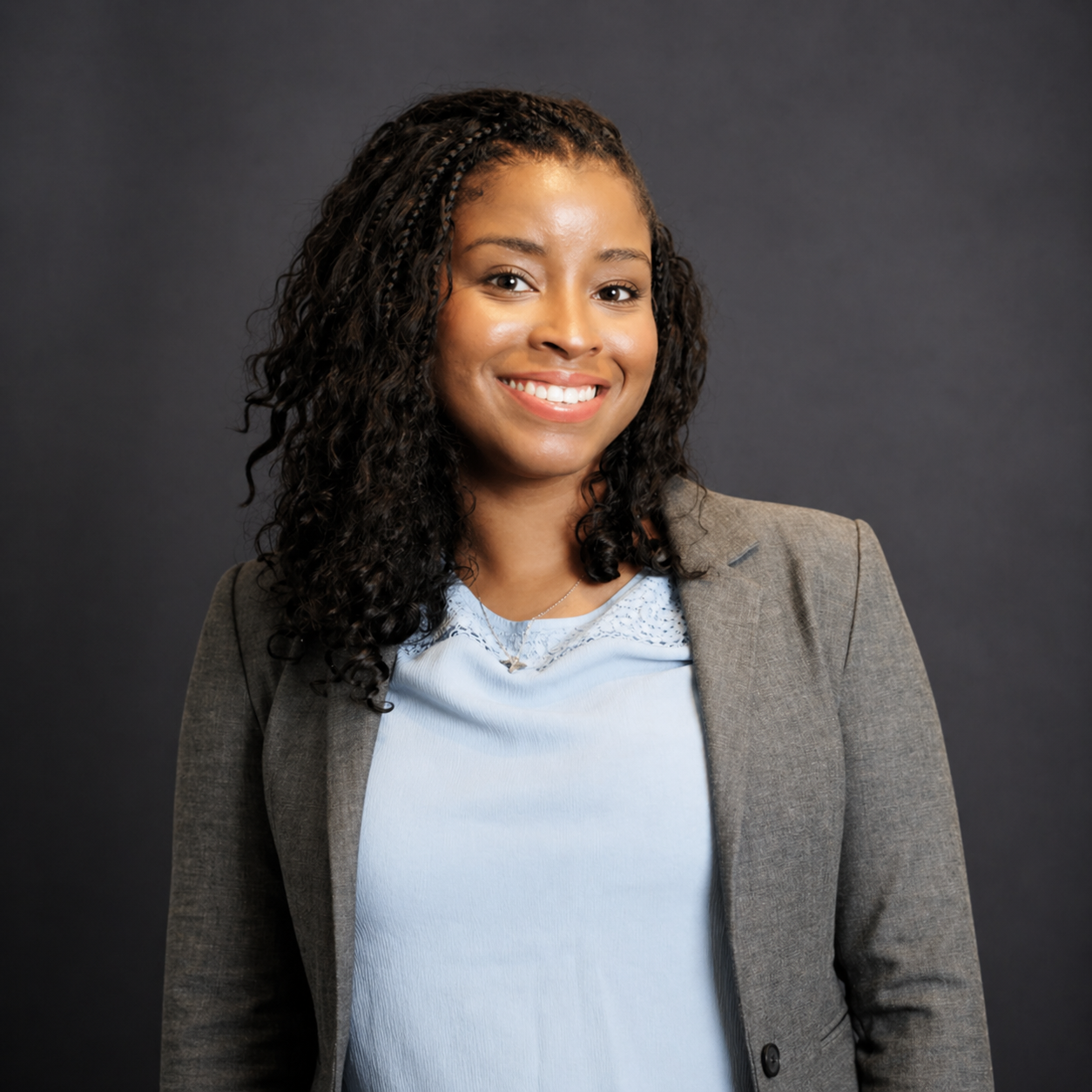 A smiling woman with curly black hair, wearing a light blue blouse and a gray blazer, standing against a dark gray background corporate professional headshot
