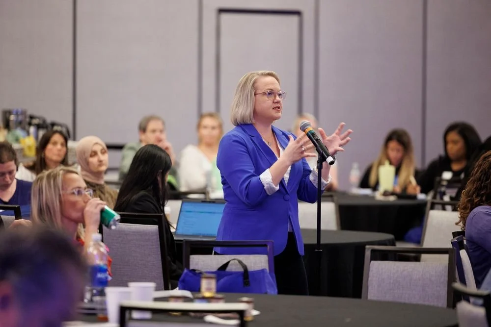 A woman in a blue blazer is speaking into a microphone during a conference or seminar, with a diverse audience seated around her.