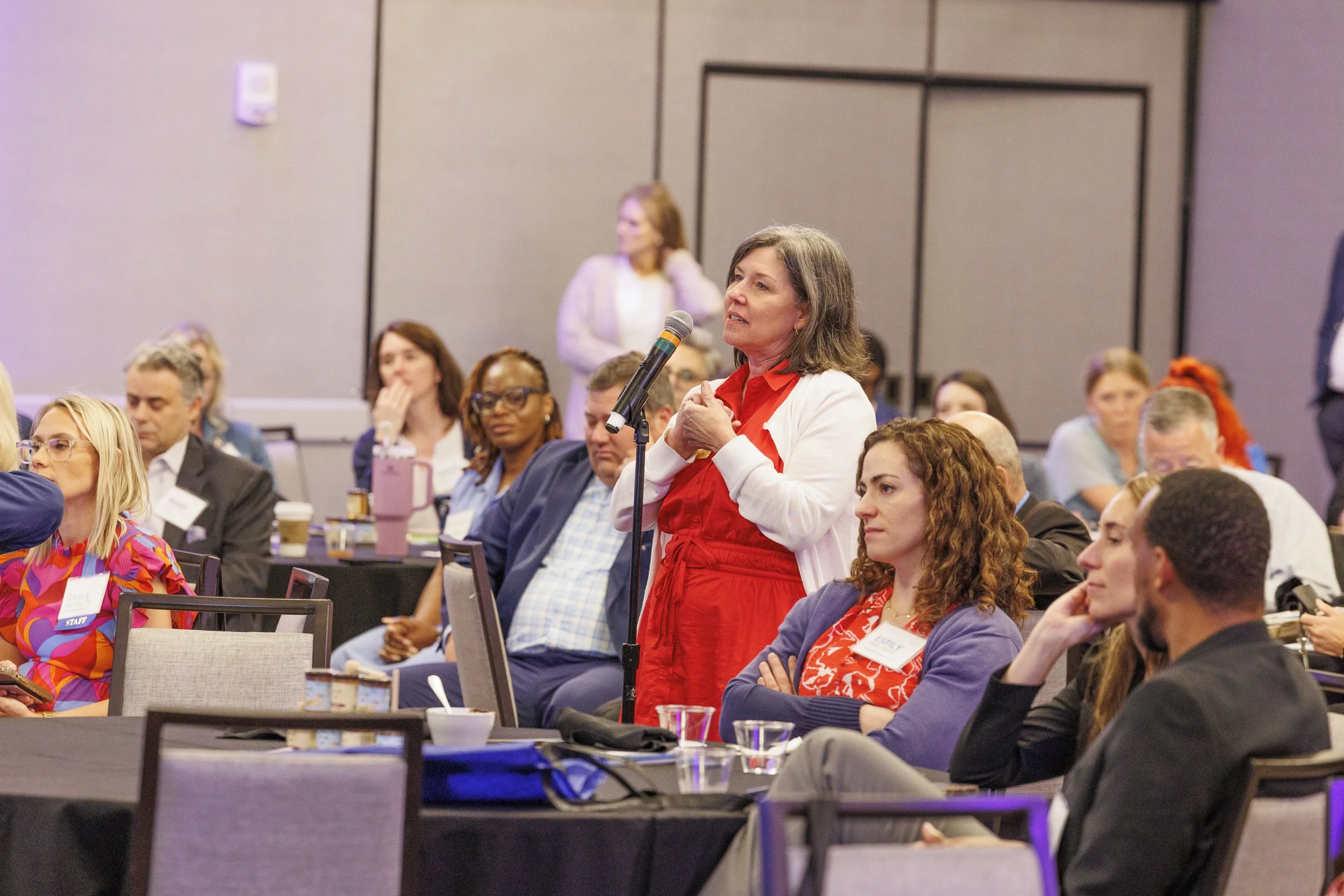 A woman in a red dress and white blazer standing at a conference microphone, speaking or asking a question, surrounded by seated attendees at a professional event.