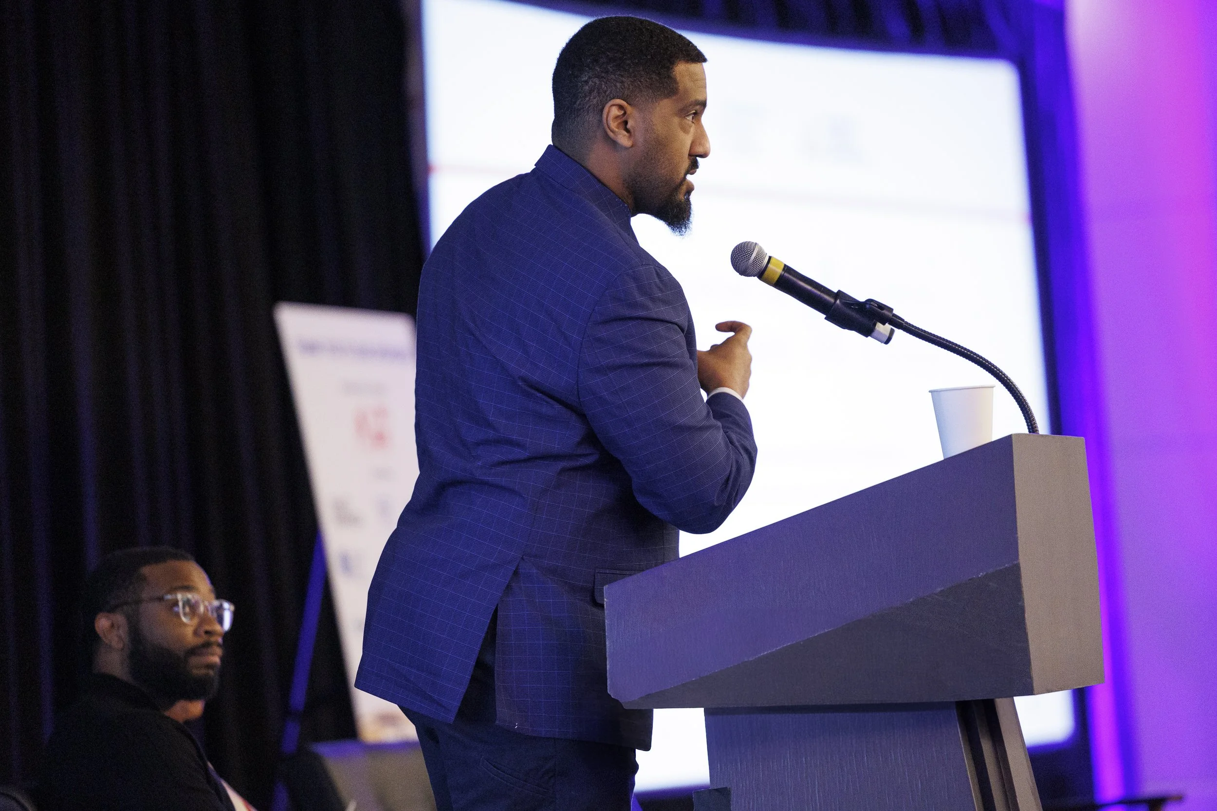 Man in a blue blazer speaking at a podium with a microphone at a conference or event.
