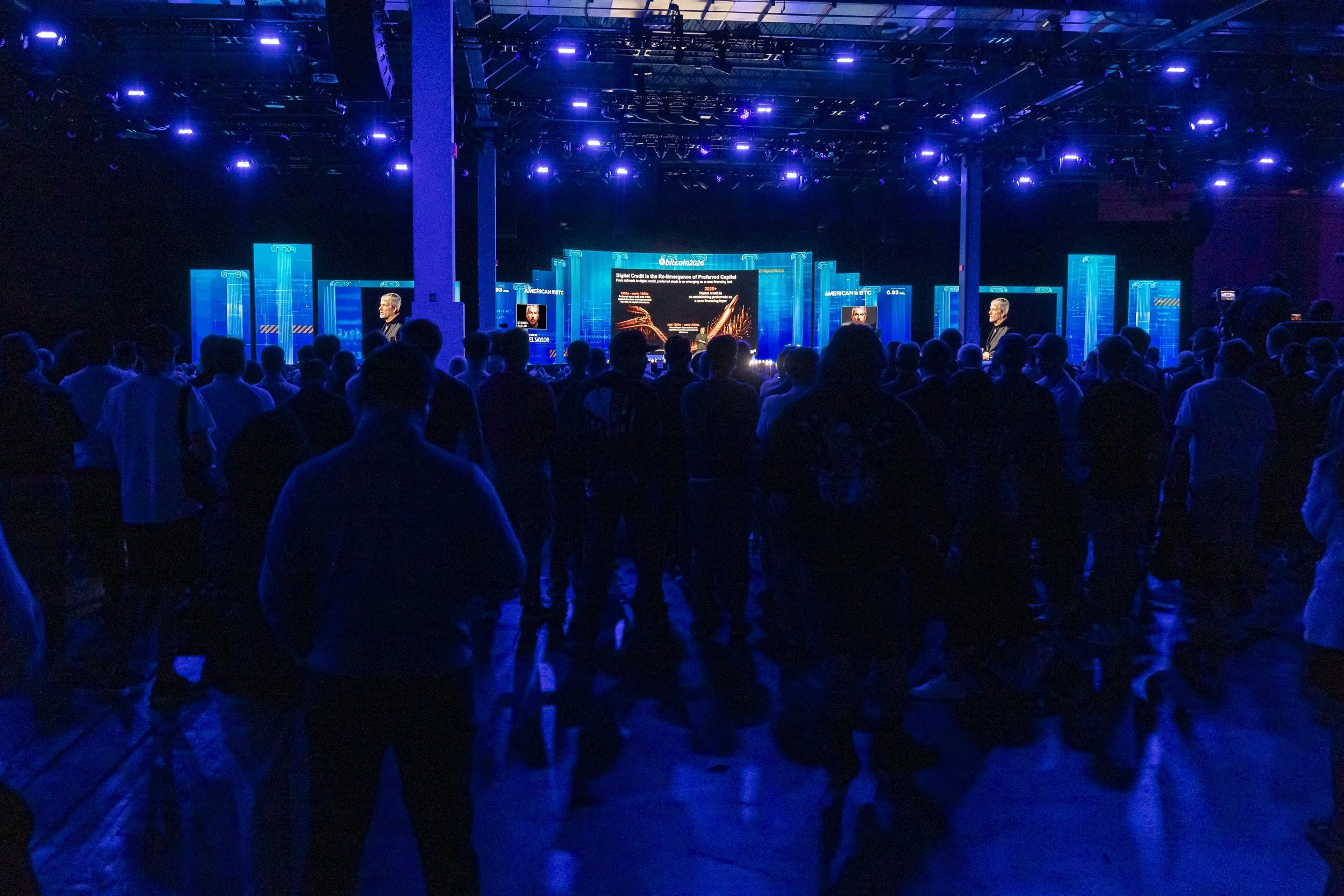 An indoor conference or event with a large audience facing a stage with multiple screens and digital displays, illuminated by blue and purple lighting.