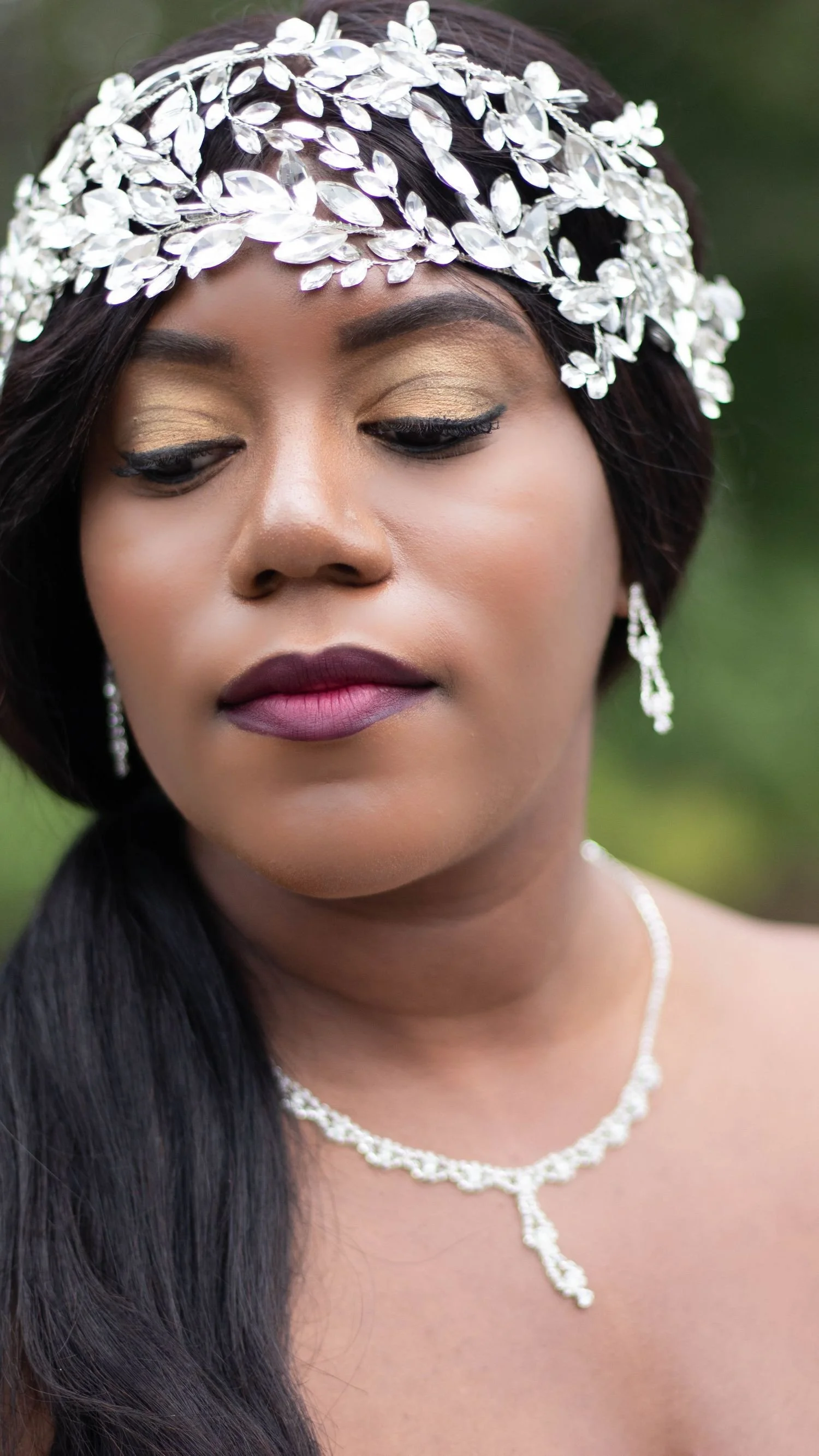 Close-up of a woman wearing a silver leaf headpiece, earrings, and a necklace with a drop pendant, with her eyes closed and dark hair styled to one side. The background is blurred green.