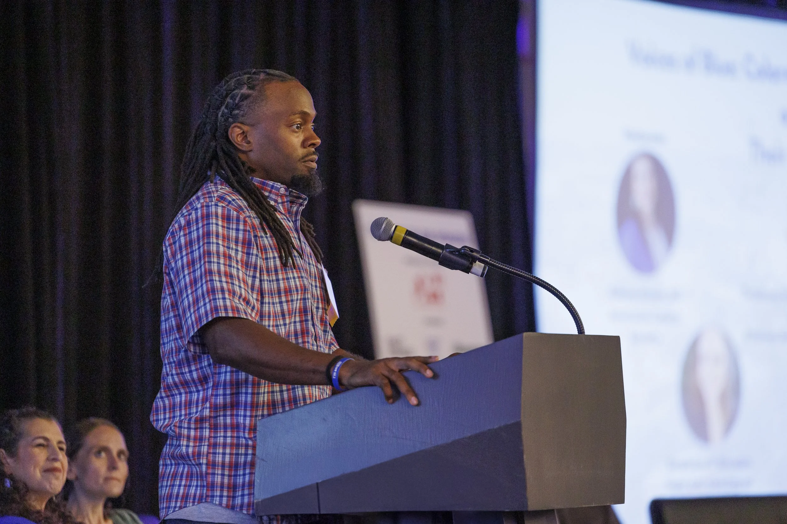 A man with long dreadlocks in a checkered shirt speaking at a podium with a microphone during a conference, with a large screen displaying a presentation in the background.