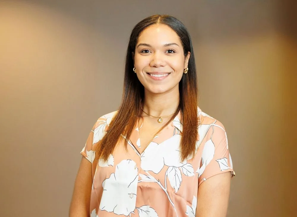 Young woman with long brown hair smiling, wearing a peach-colored floral blouse and jewelry, standing against a plain background.