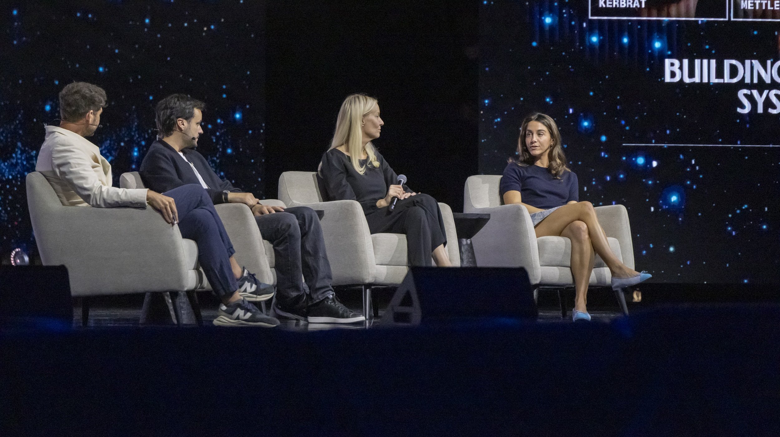 Four people sitting on stage in chairs during a panel discussion at a conference, with a digital background and a partial view of text on a screen.