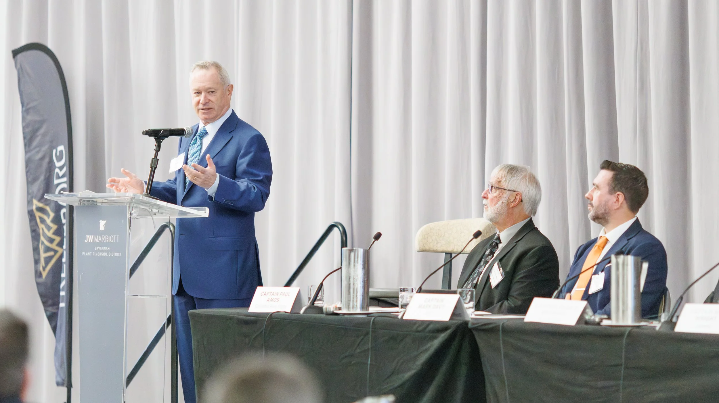 A man in a blue suit is speaking at a podium with a microphone, while two men sit at a table with microphones and nameplates, in a conference room with white curtains in the background.