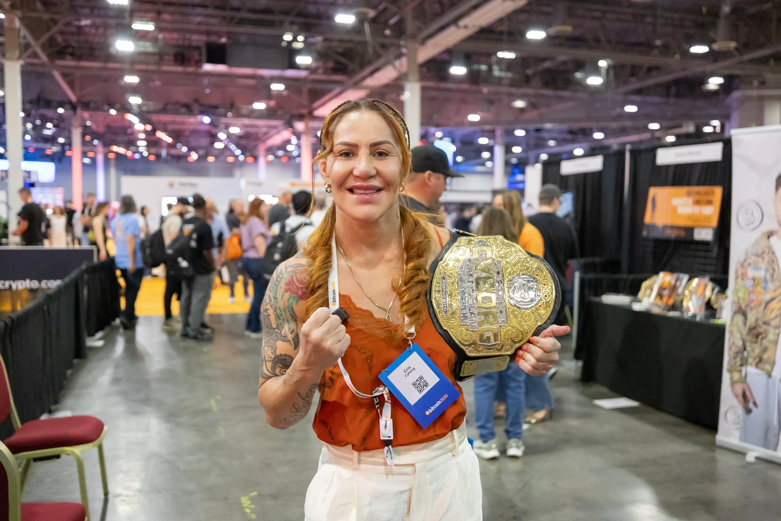A woman with red hair, tattoos, and earrings holding a large gold championship belt at a crowded indoor event.