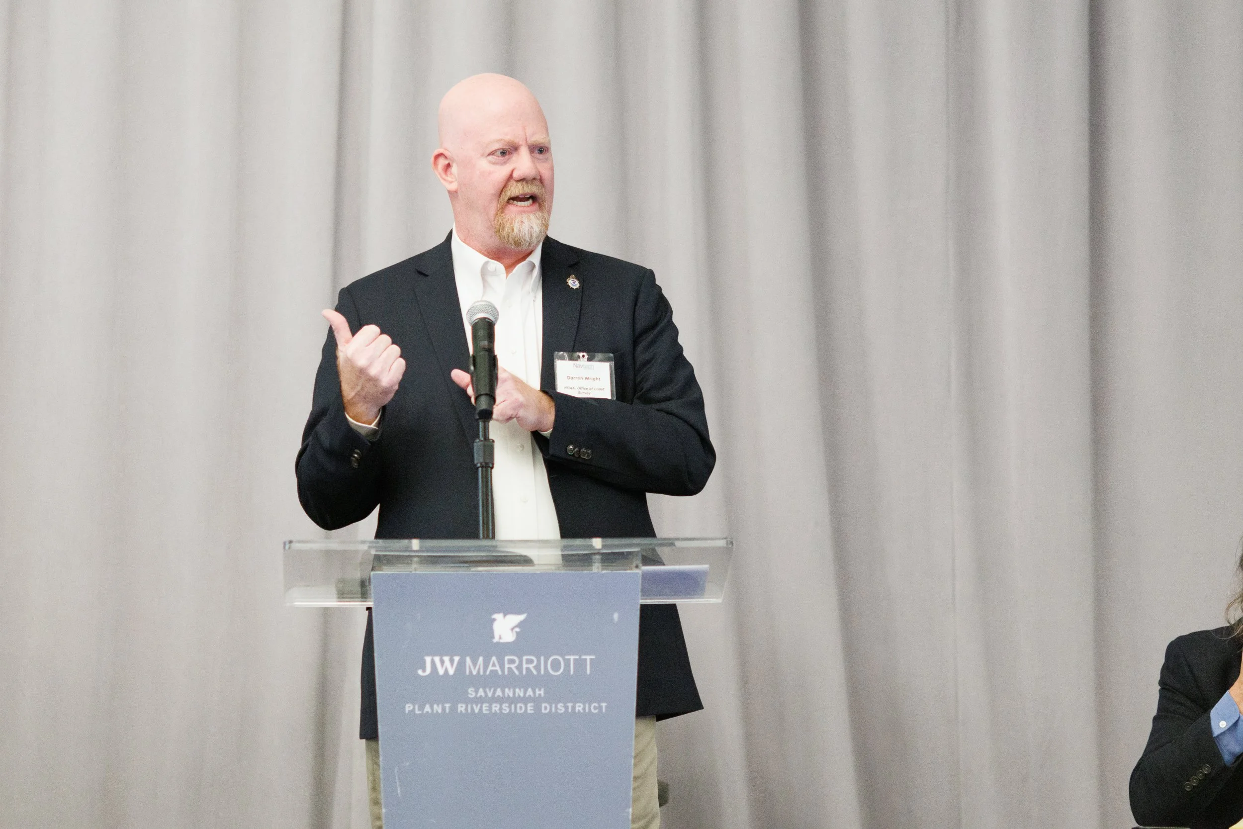 A man with a beard and bald head, wearing a black suit and white shirt, speaking at a podium with a microphone at the JW Marriott Savannah Plant Riverside District hotel.