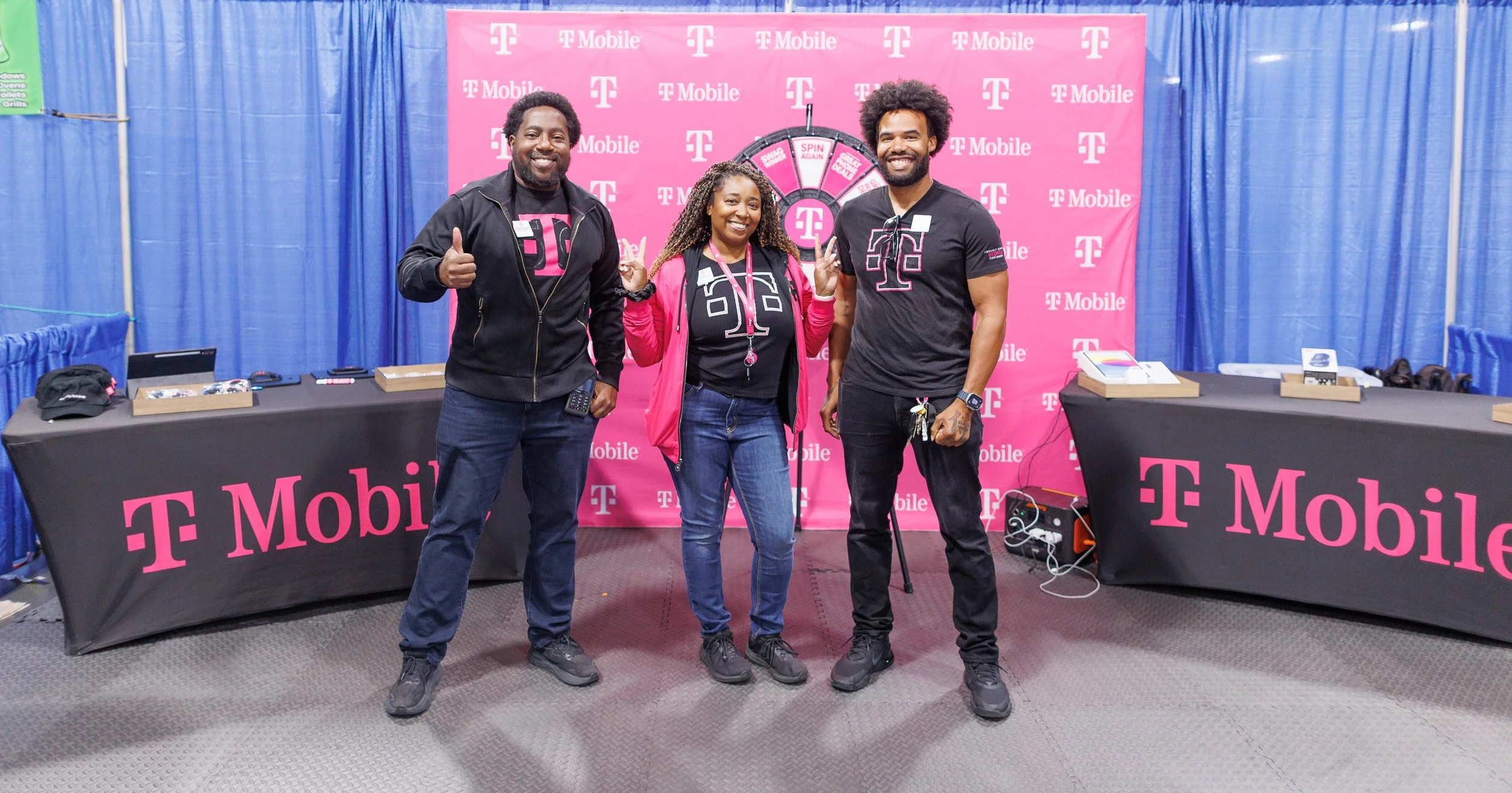 Three people standing in front of a pink T-Mobile backdrop with a spinning wheel game behind them at a T-Mobile event. They are smiling and making peace signs.