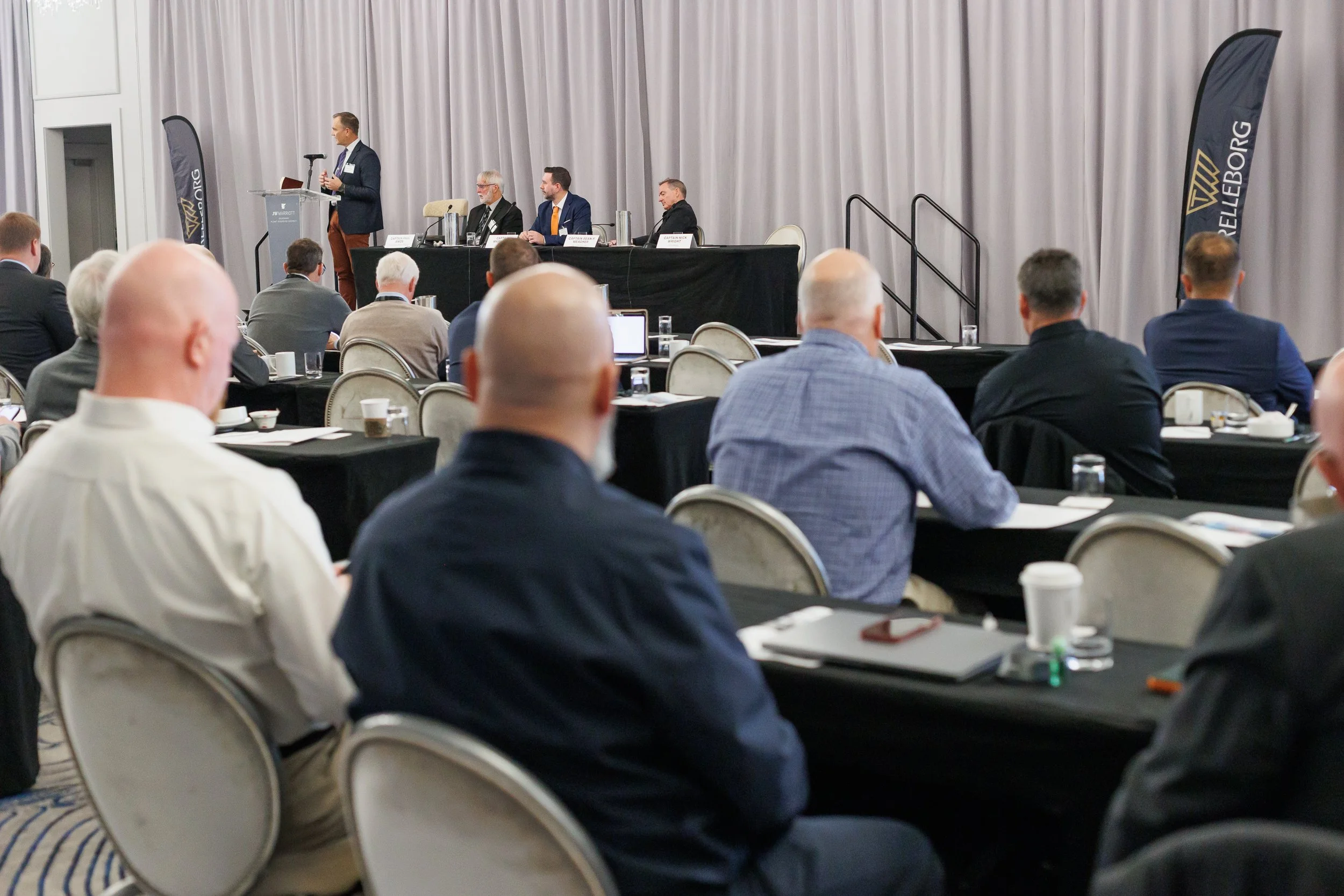 A man speaking at a podium during a conference, with four seated panelists on stage and an audience listening, in a large conference room with grey curtains.