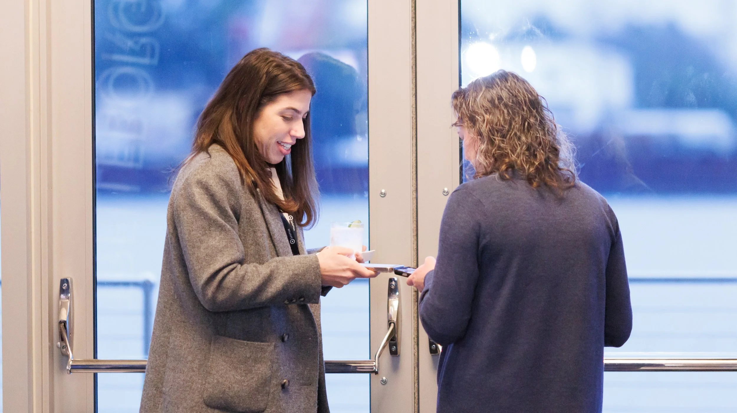 Two women standing by glass doors, one handing the other a phone, both smiling, during daytime.