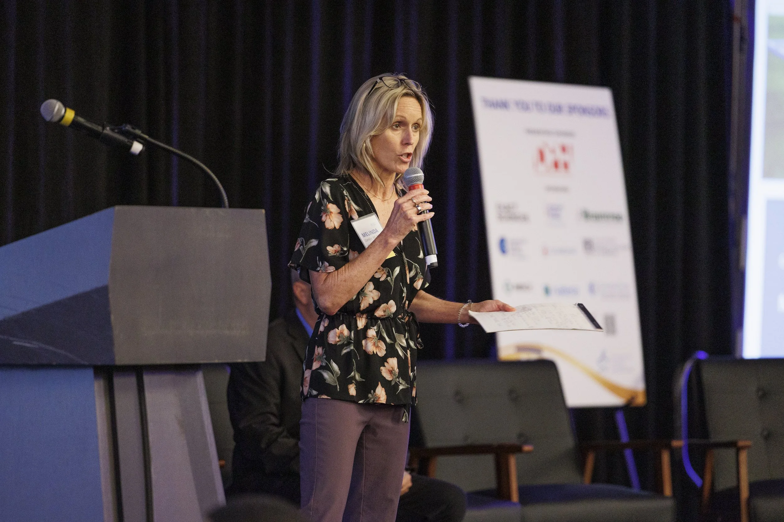 A woman with blonde hair speaking at a conference, holding a microphone and a sheet of paper, standing next to a podium.