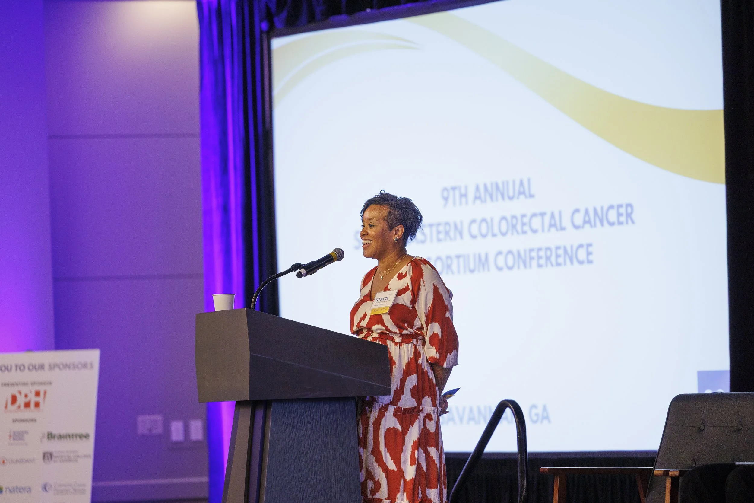 A woman in a red and white patterned dress speaking at a podium during the 9th Annual Western Colorectal Cancer Summit Conference in Savannah, Georgia.