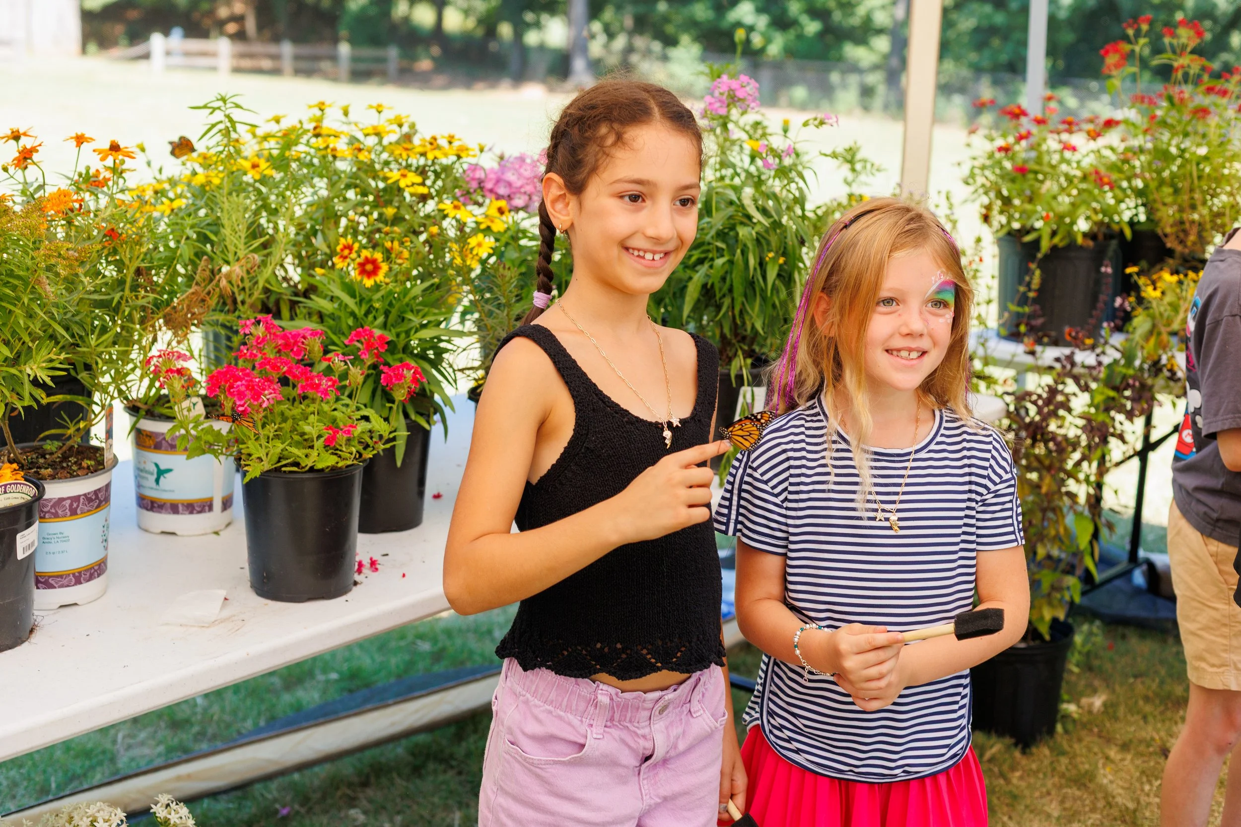 Two young girls standing in front of a table with potted flowers, outdoors during daytime. One girl is smiling and has brown hair in a braid, wearing a black top and pink shorts. The other girl has blonde hair, colorful face paint, and is wearing a s