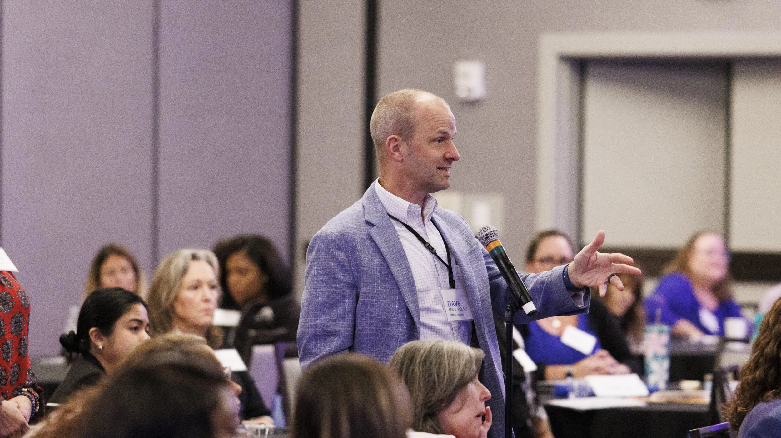 A man in a light purple blazer and checkered shirt speaking into a microphone during a conference. He is standing and gesturing with his right hand, with several seated women listening in the background.