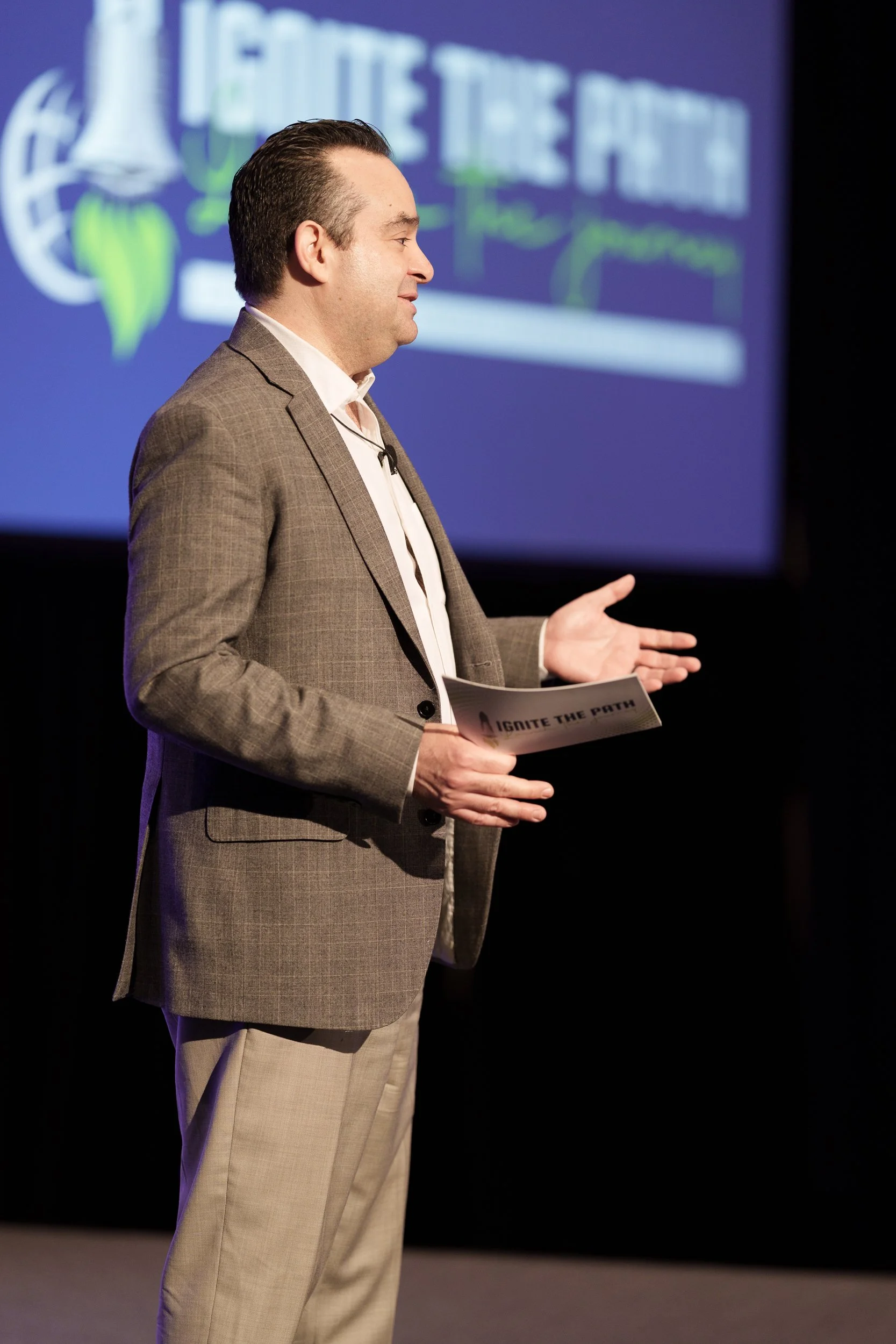 Man in plaid suit giving a presentation, holding a paper titled 'Ignite the Path,' with a blue background & a blurred slide in the background at the Westin Peachtree Plaza; corporate photography cover also covers JW Marriott Savannah Plant Riverside