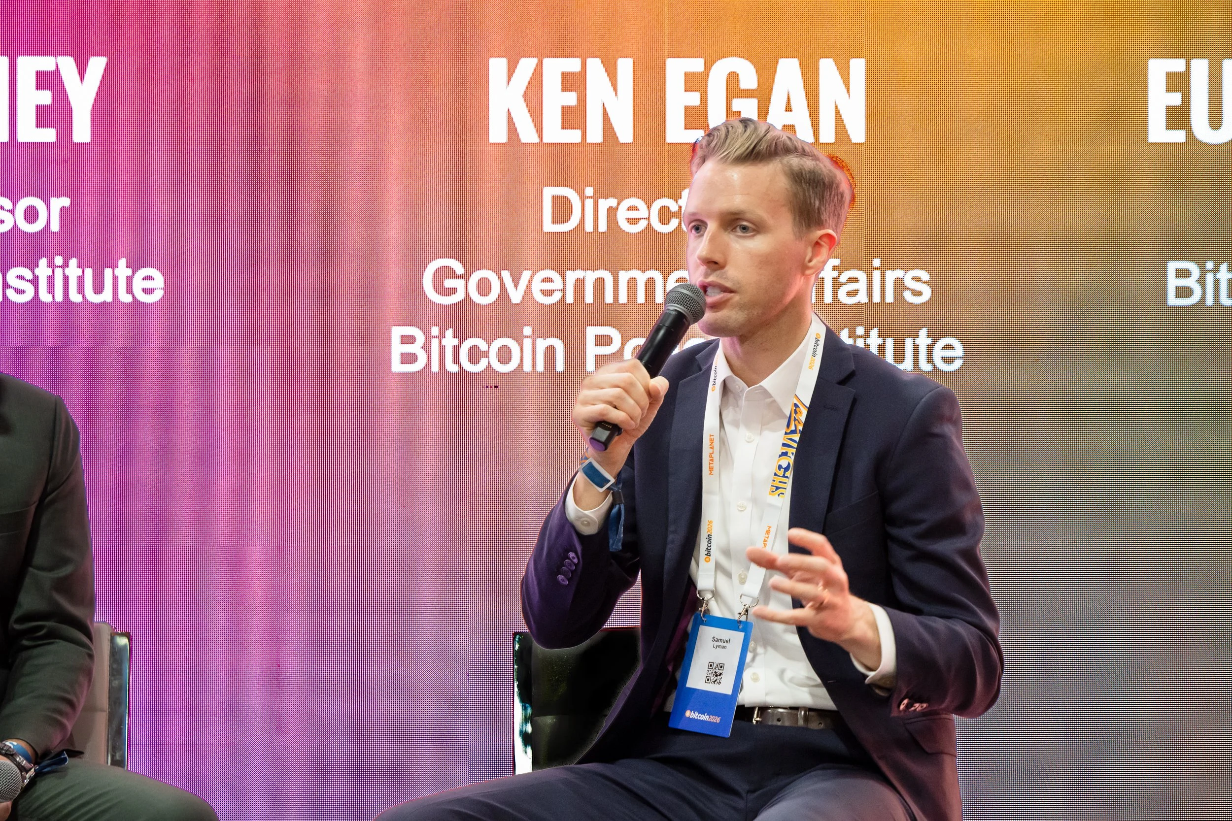 A man in a suit speaking into a microphone at a conference or panel discussion, with a colorful background displaying text related to government and cryptocurrency.