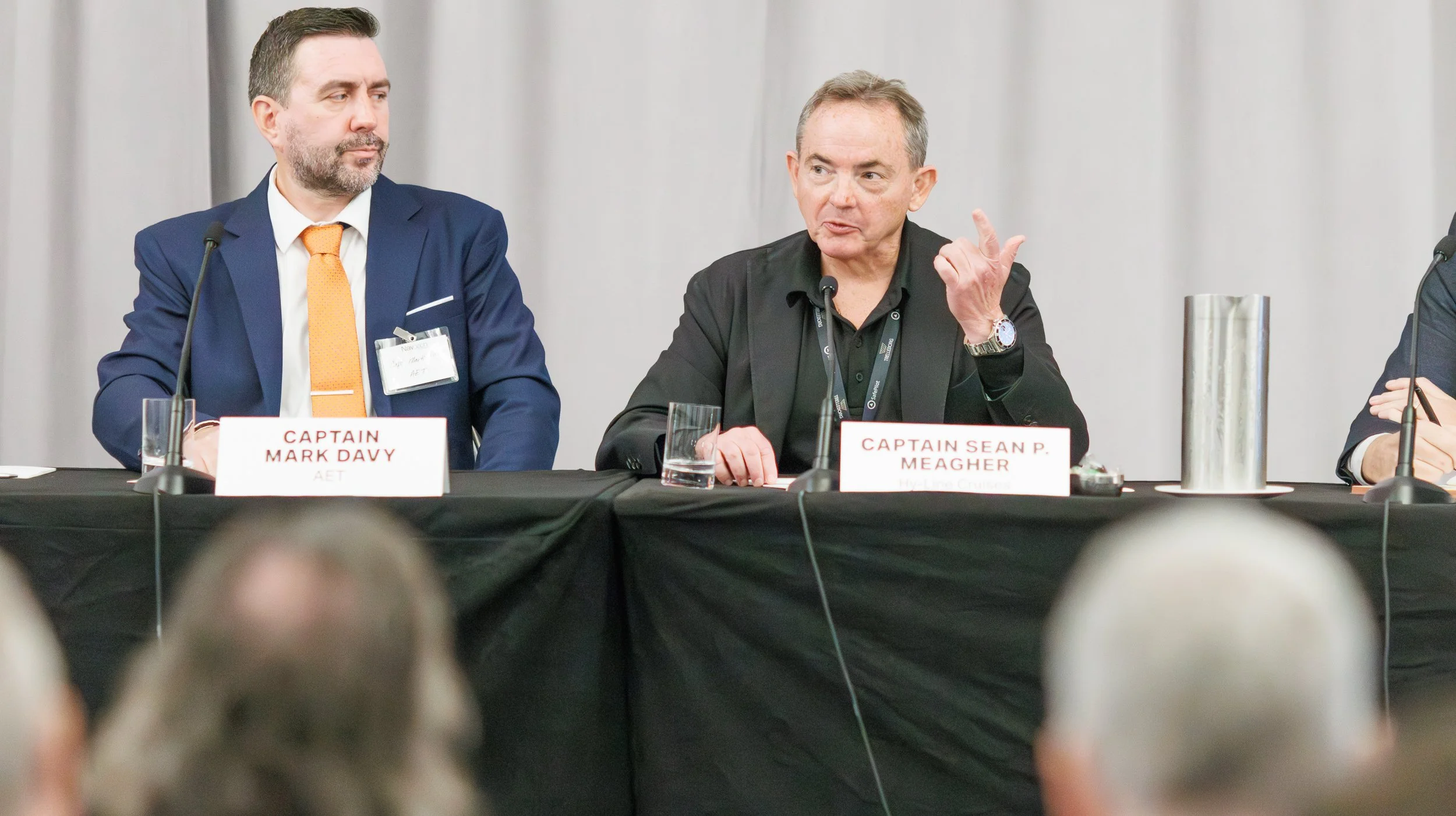 Two men sitting at a conference table with nameplates. The man on the left, wearing a blue suit and orange tie, has a nameplate reading 'Captain Mark Davy'. The man on the right, in a black shirt, has a nameplate reading 'Captain Sean P. Meagher'; he