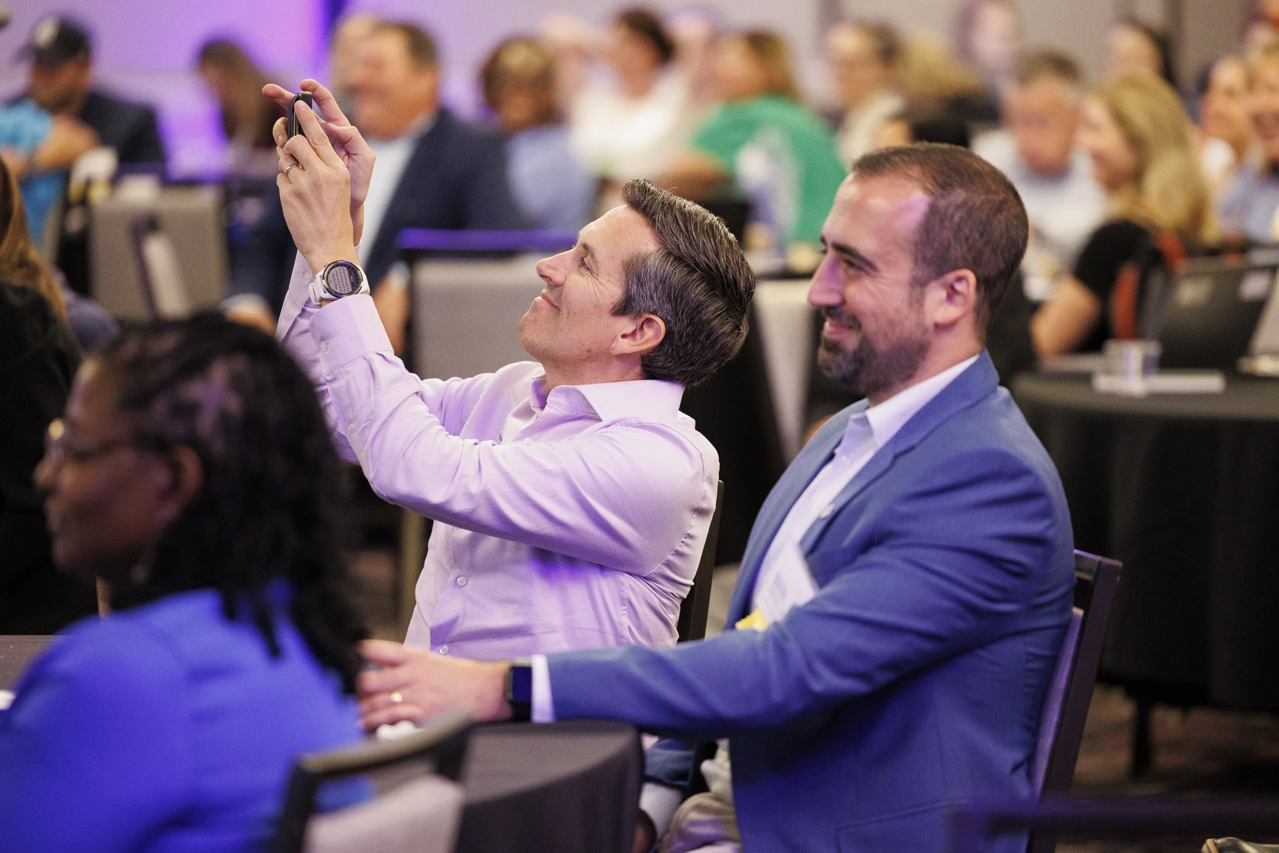 Two men sitting at a conference, one taking a photo with his phone, and the other smiling, surrounded by other attendees.