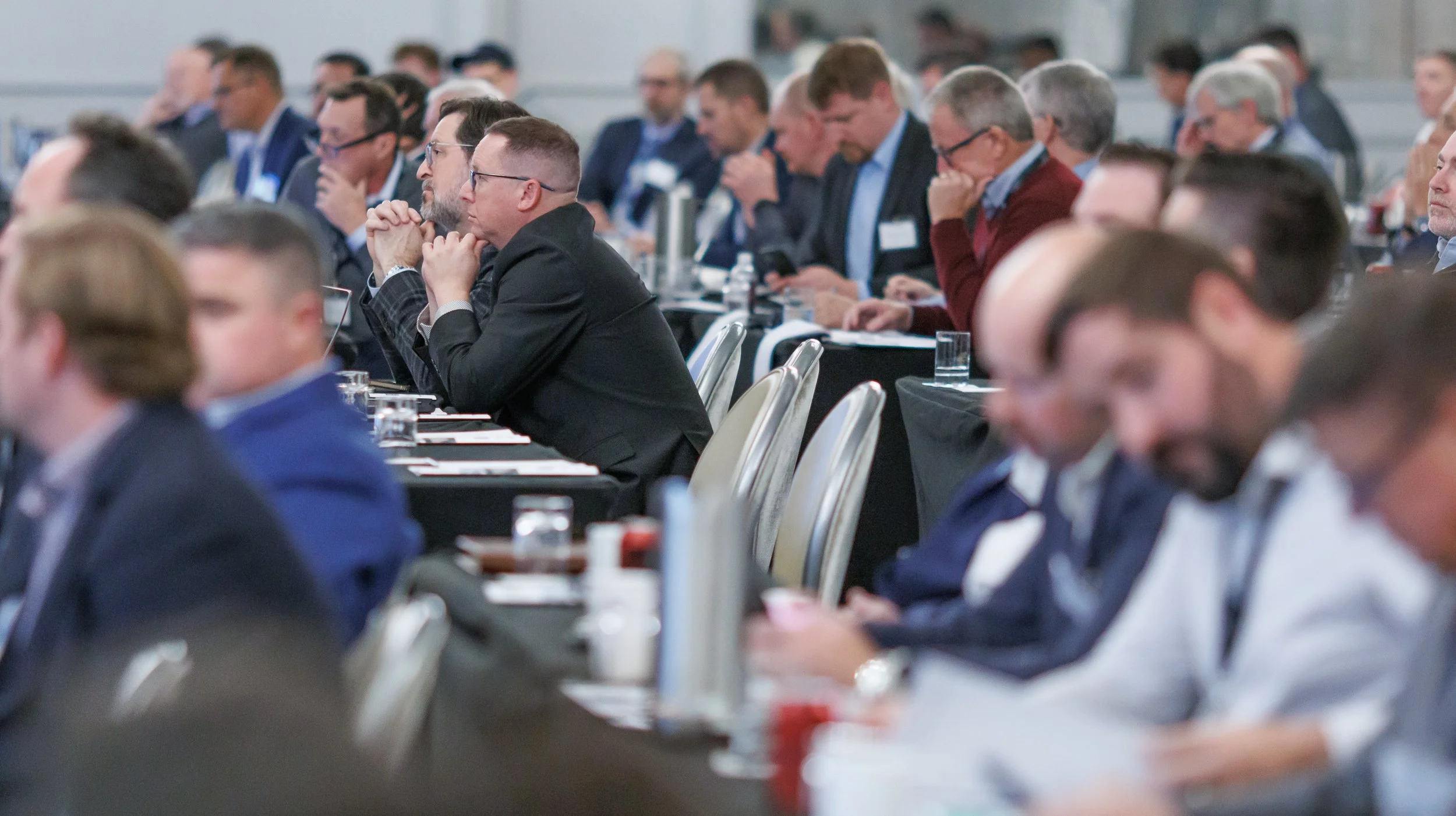 Attendees at a professional conference or seminar sitting at tables, reading, listening, and taking notes.