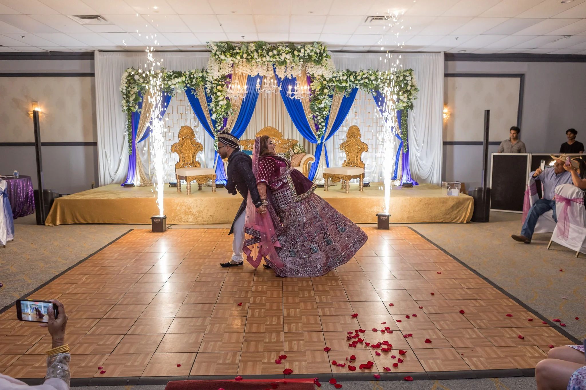 A bride and groom dancing at their wedding reception on a decorated stage with blue and white drapery, floral arrangements, golden chairs, and sparklers on the dance floor, with guests seated around watching.