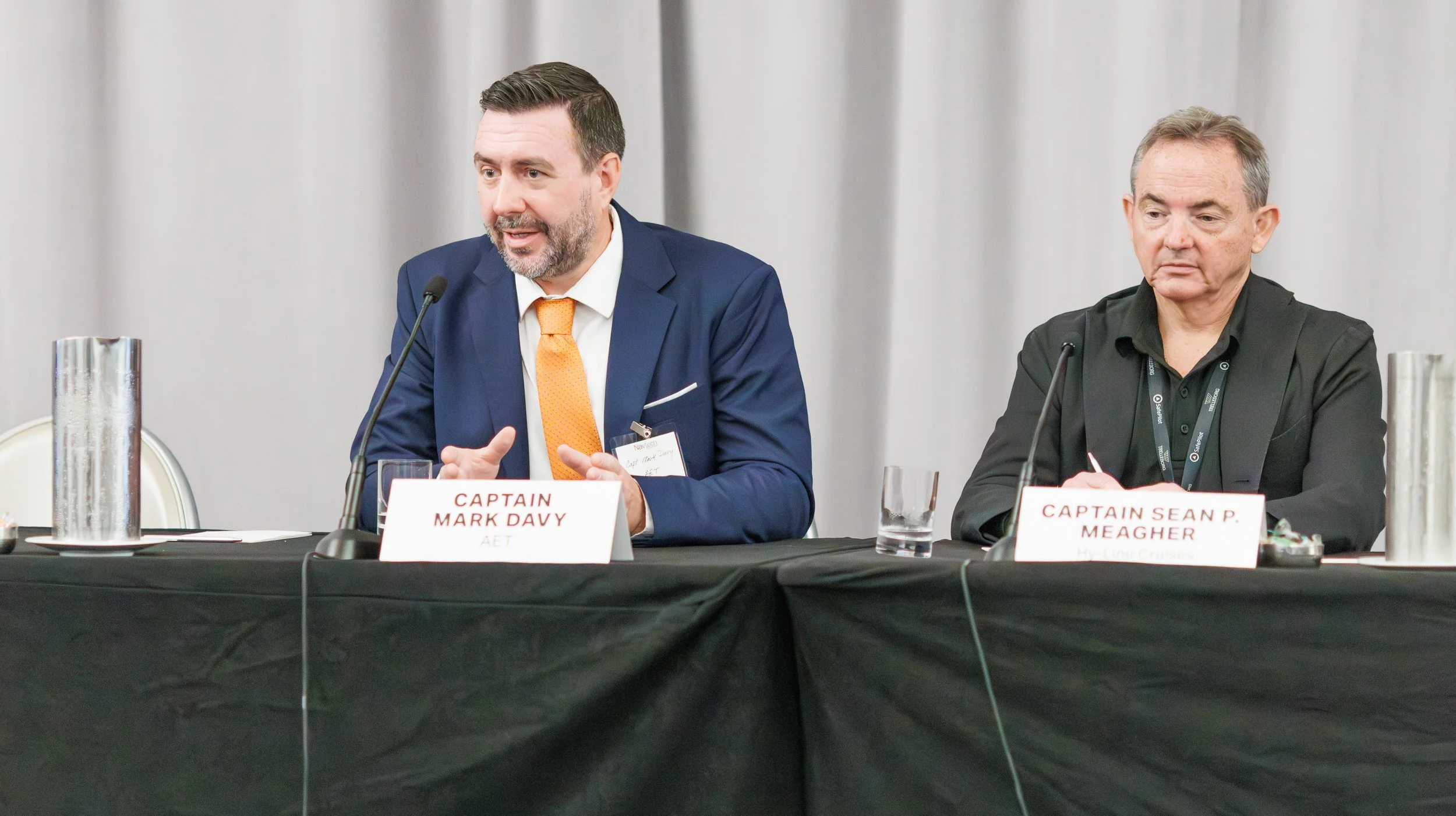 Two men sitting at a conference table speaking into microphones. The man on the left, wearing a blue suit and orange tie, is identified as Captain Mark Davy, and the man on the right, in a black shirt, is Captain Sean P. Meagher. There are nameplates