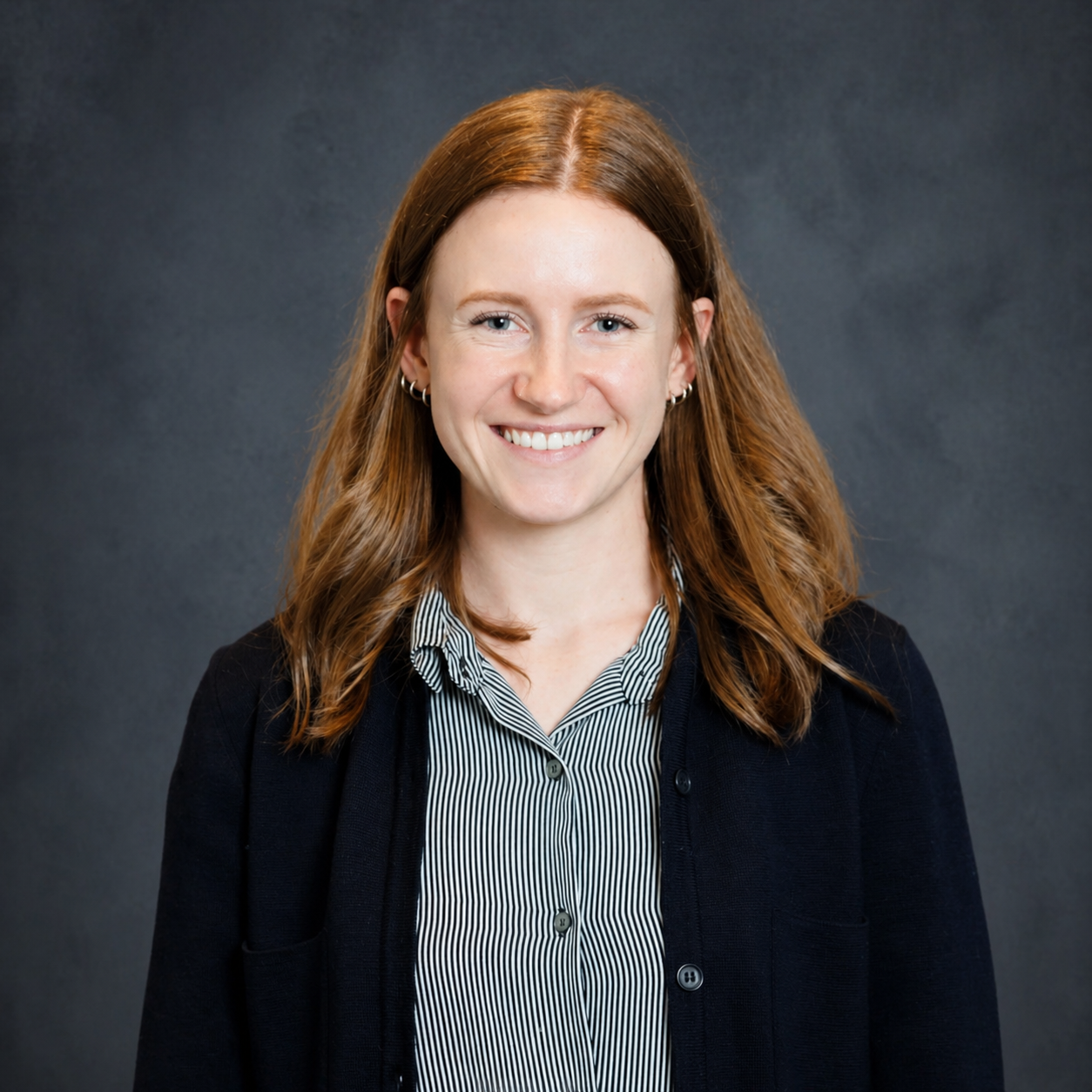 A young woman with red hair, smiling, wearing a striped shirt and dark cardigan, against a dark gray background. corporate professional headshot