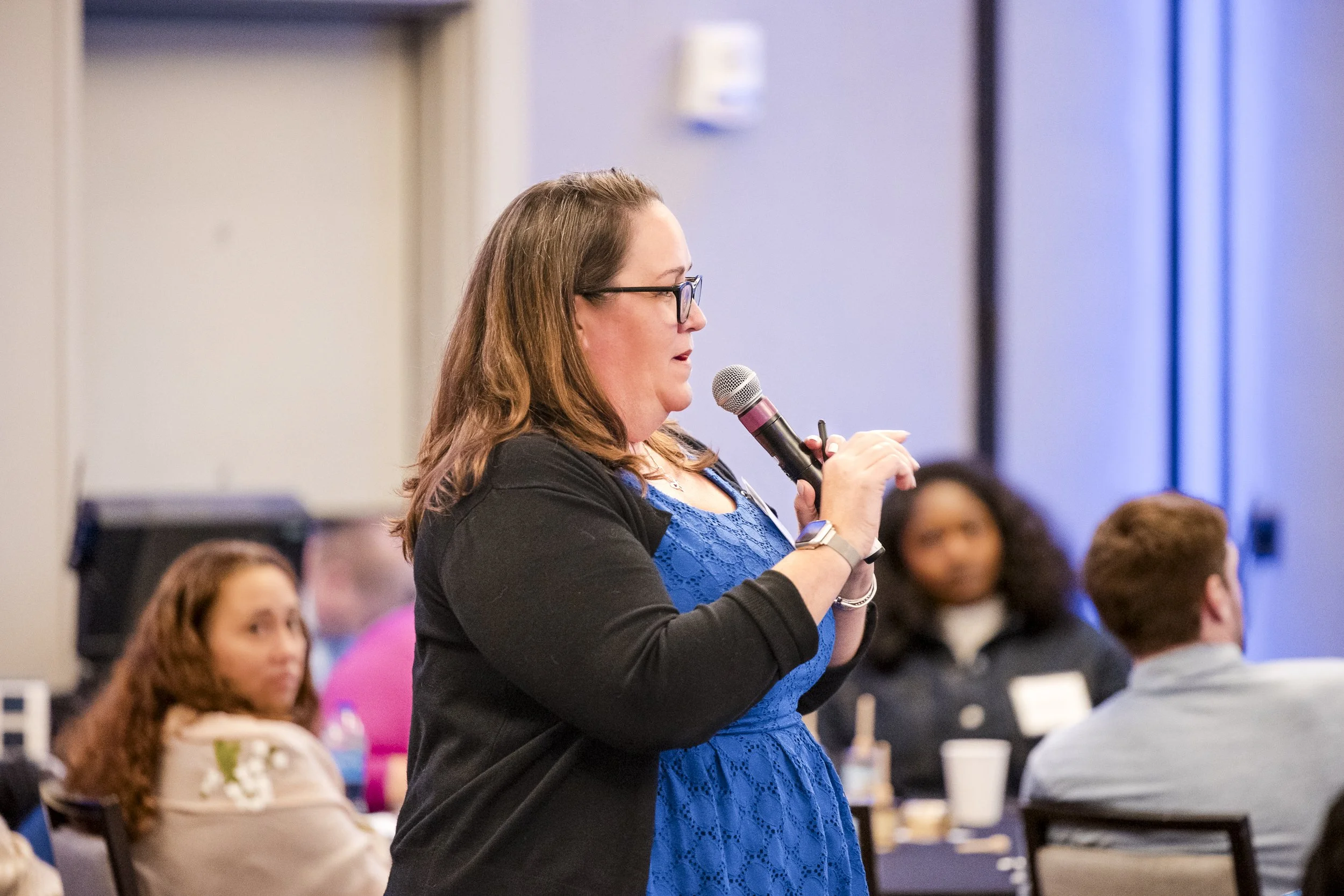 A woman with glasses and brown hair holding a microphone, speaking at an indoor event with people seated at tables in the background.
