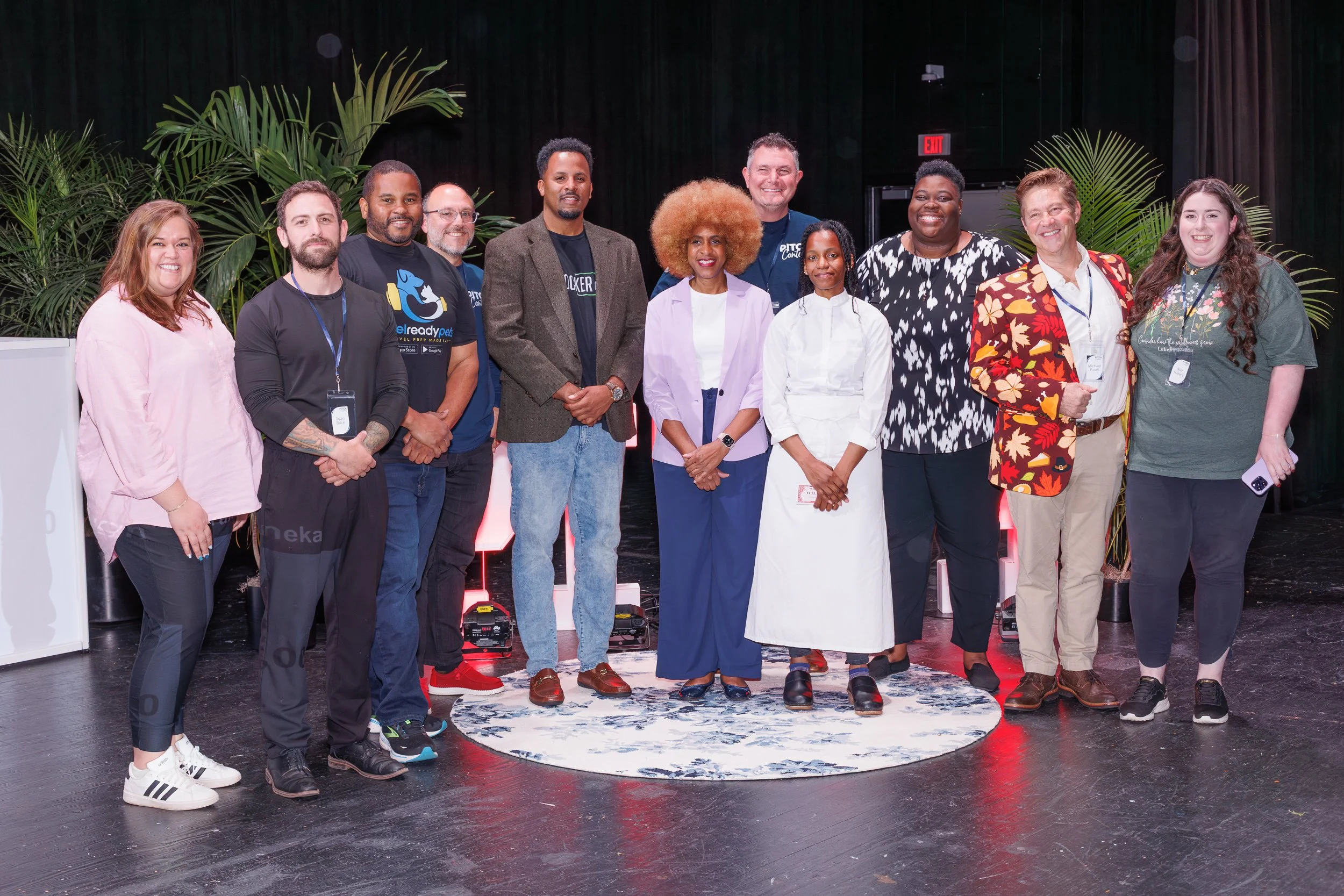 A diverse group of twelve people standing together on a stage with a black backdrop and green plants. Some are wearing casual clothing, others business attire, and they are smiling for the photo.