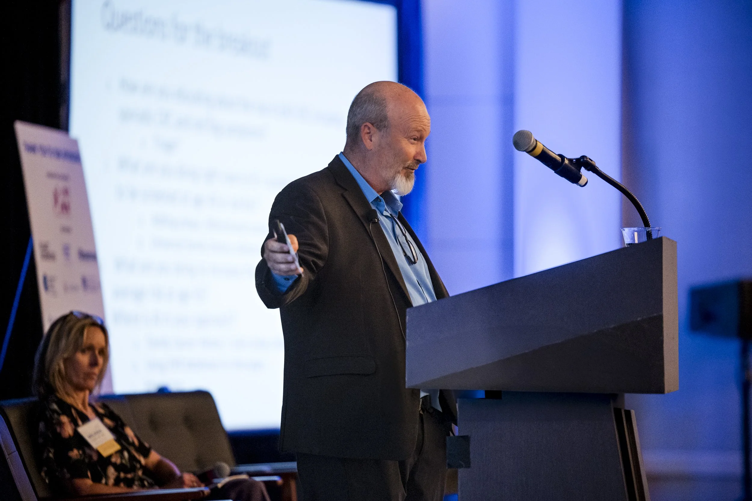 Man giving a presentation at a conference, standing behind a podium with a microphone, gesturing with his hand, with a woman seated in the background.