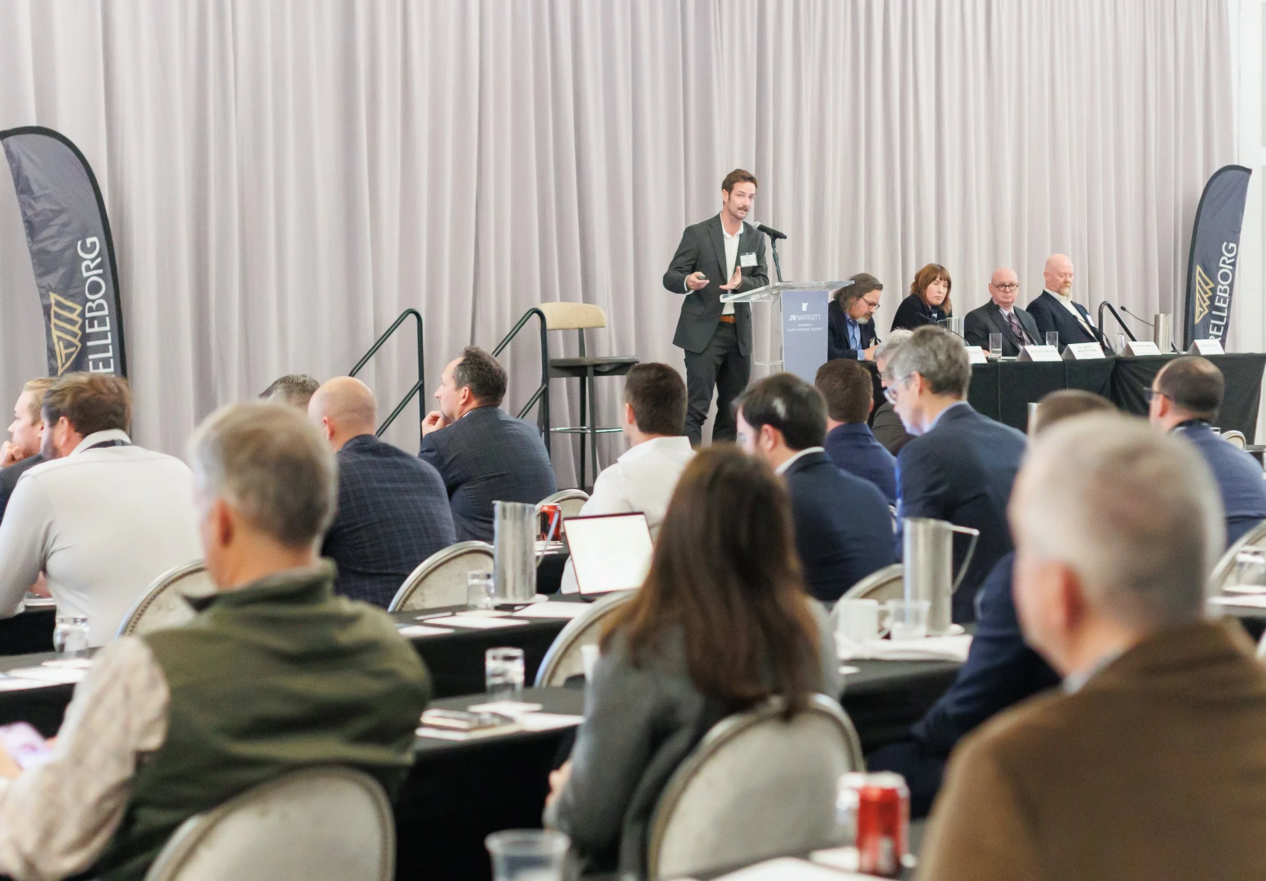 A businessman in a dark suit giving a presentation at a conference, with a panel of four seated professionals behind him, and an audience of mostly businesspeople listening in a conference room with white curtains and banners.