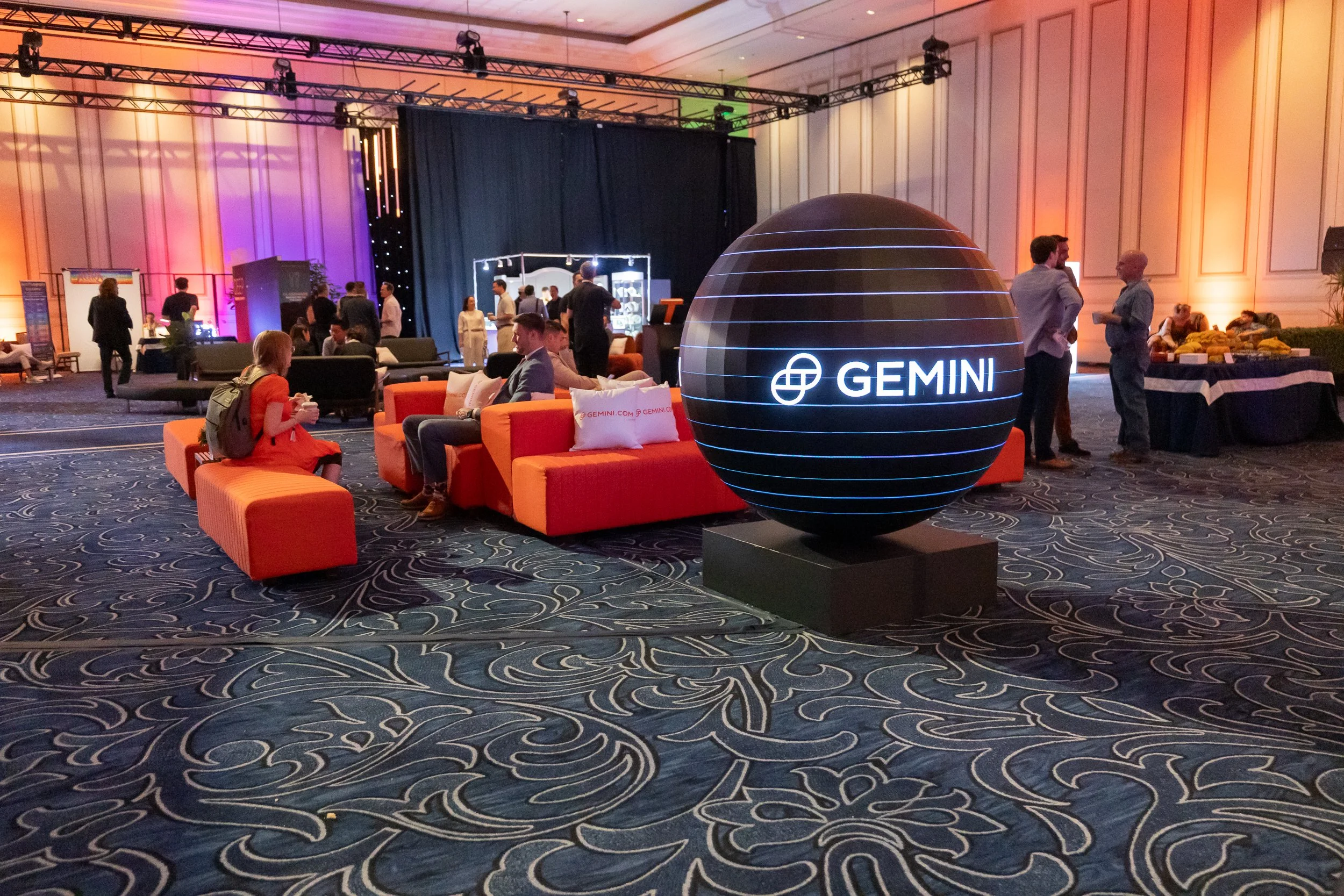 A conference or event space filled with people, with a large illuminated Gemini logo globe in the foreground. The room has colorful lighting on the walls, seating areas with orange and dark-colored furniture, and a stage setup with black curtains and