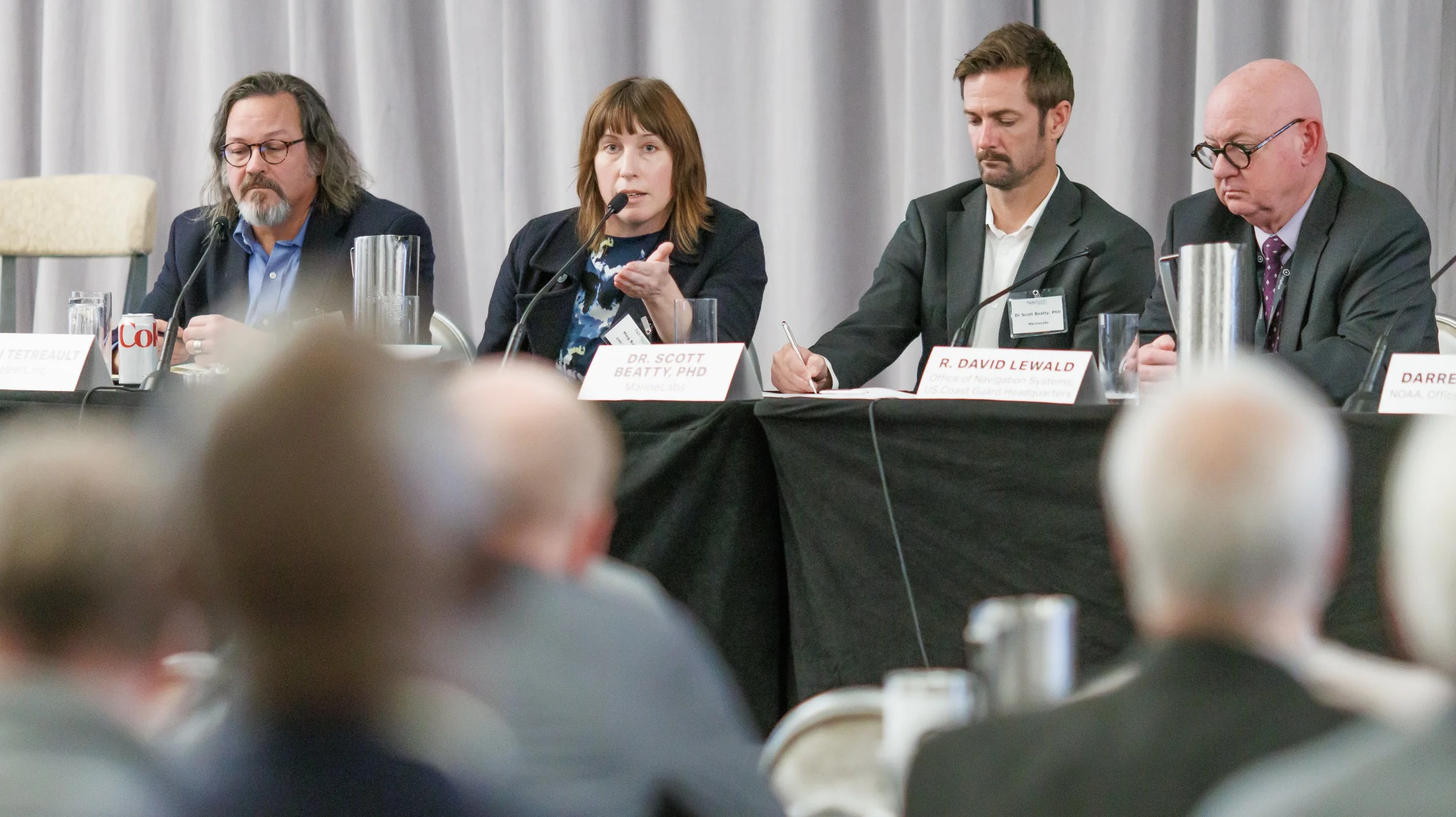 A panel of four people sitting at a long table during a conference or meeting. One woman is speaking into a microphone, while the others are listening or taking notes. There are nameplates and microphones in front of each person, with an audience in 