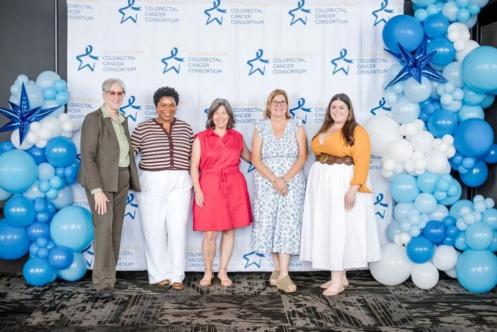 Five women standing together at a Southeastern Colorectal Cancer Consortium event, with a backdrop and balloon decorations in shades of blue and white.