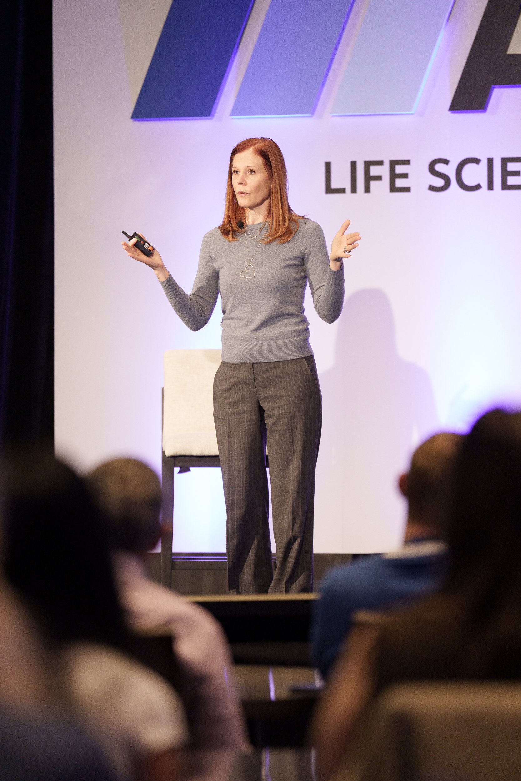 A woman with long red hair wearing a gray sweater and plaid pants giving a presentation in front of a large screen with the words 'LIFE SCI' visible.