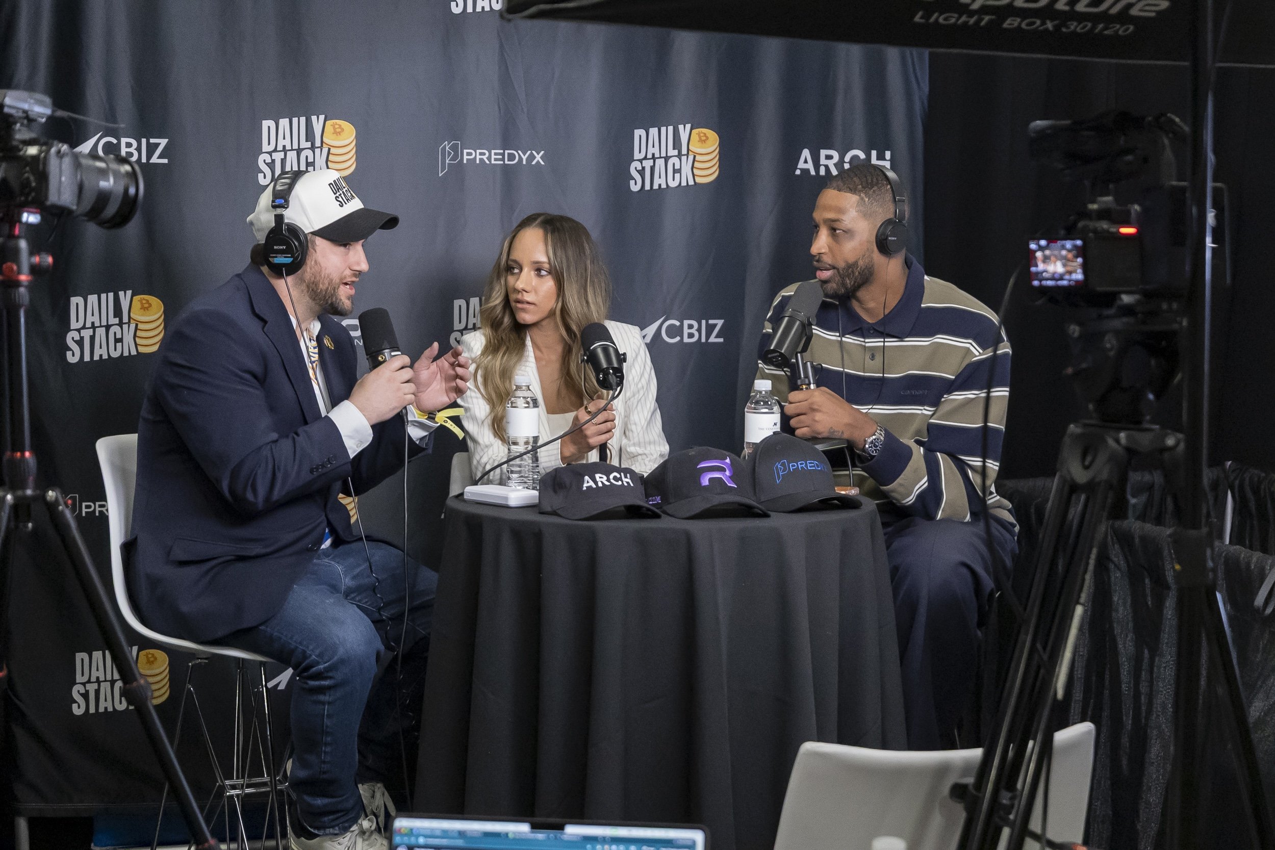 Three people sitting at a table during a podcast interview, with a backdrop displaying the words 'Daily Stack' and sponsor logos, including 'PredyX' and 'ArcH,' with recording equipment and cameras nearby.