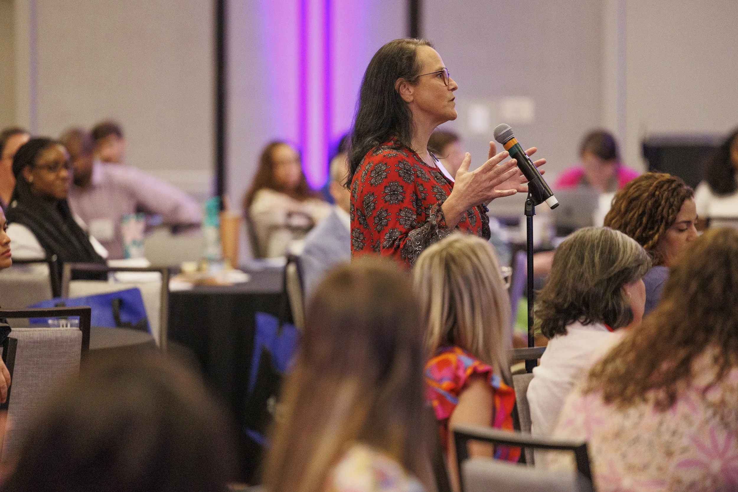A woman in a red patterned blouse stands and speaks into a microphone at a conference or seminar, surrounded by seated attendees.