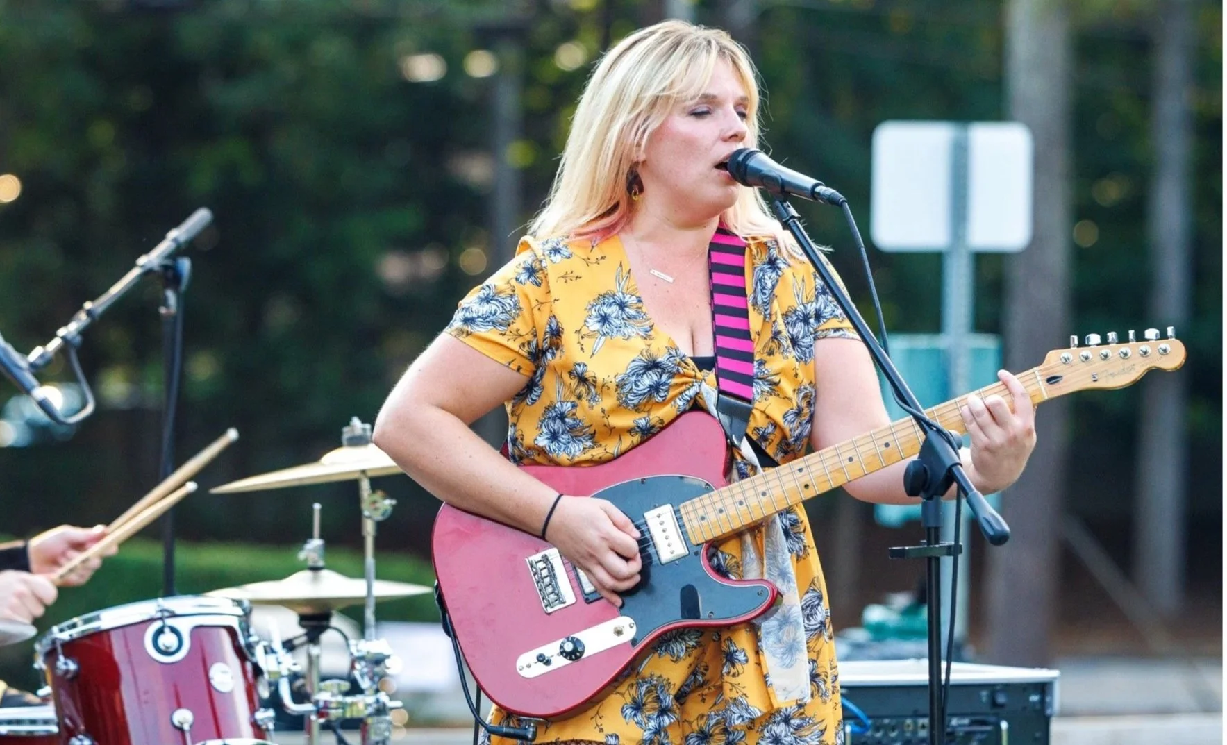 A woman with blonde hair, wearing a yellow floral dress, singing into a microphone while playing an electric guitar with a pink body during an outdoor performance.