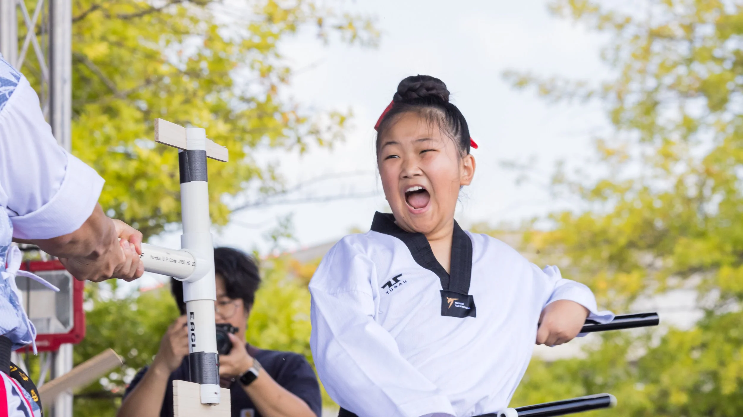 A young girl wearing a taekwondo uniform smiling and celebrating outdoors, beside a pole with a cross-shaped top, with trees in the background.
