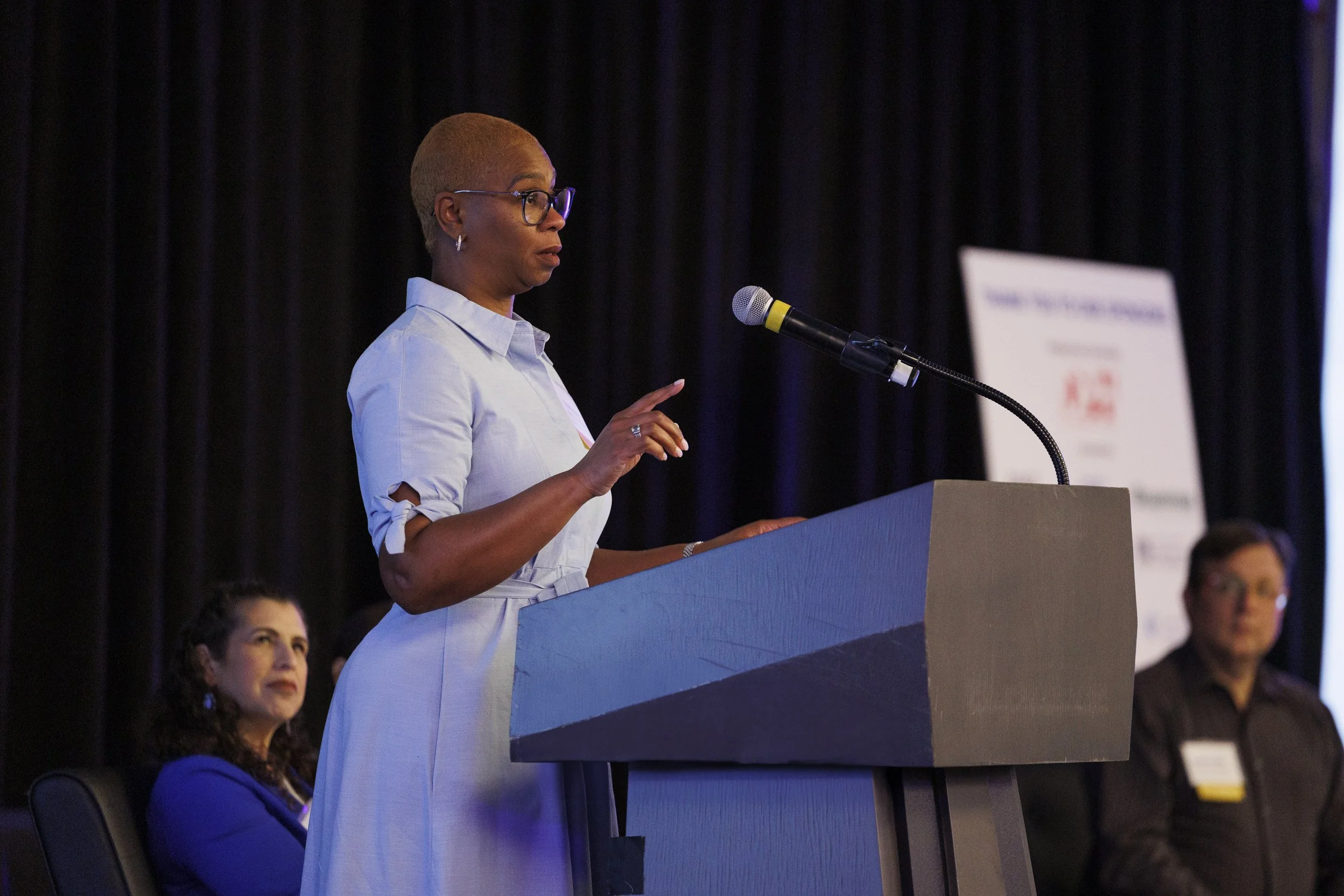 A woman in glasses, wearing a light blue dress, is speaking at a podium during an event or conference, with a dark curtain background. Other people are seated behind her, including a woman in a blue blazer and a man in a black shirt.