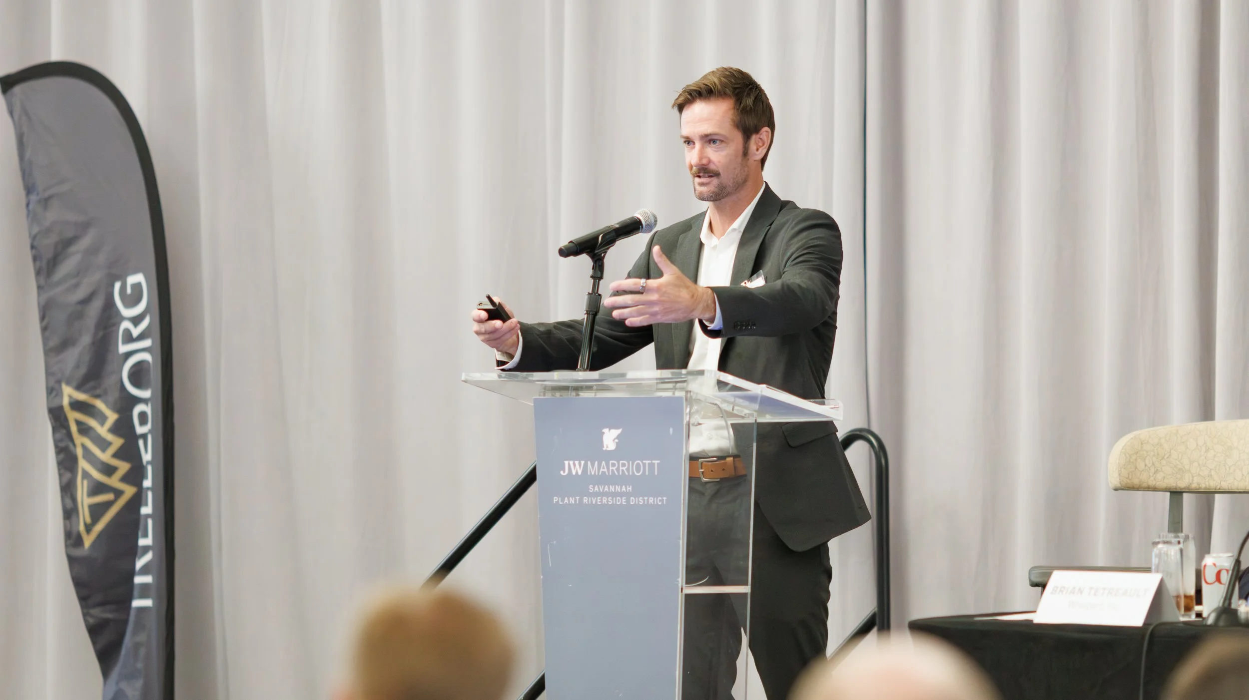 A man in a dark suit giving a presentation at a conference held at JW Marriott in Savannah, standing behind a clear podium with a microphone, gesturing with one hand while holding a remote in the other, with a banner and a table with a name card visi