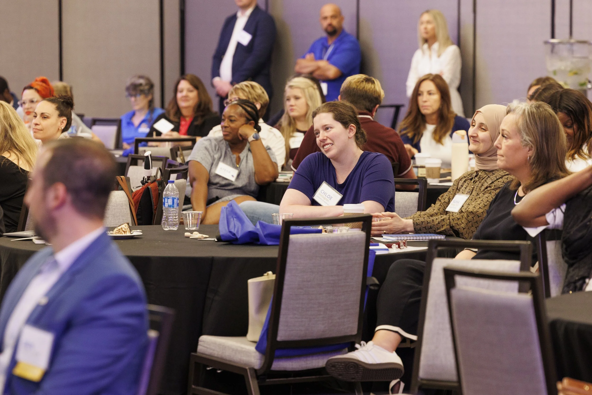 A diverse group of women and men attending a conference or seminar, seated at round tables with notebooks, drinks, and name tags, listening attentively and smiling.