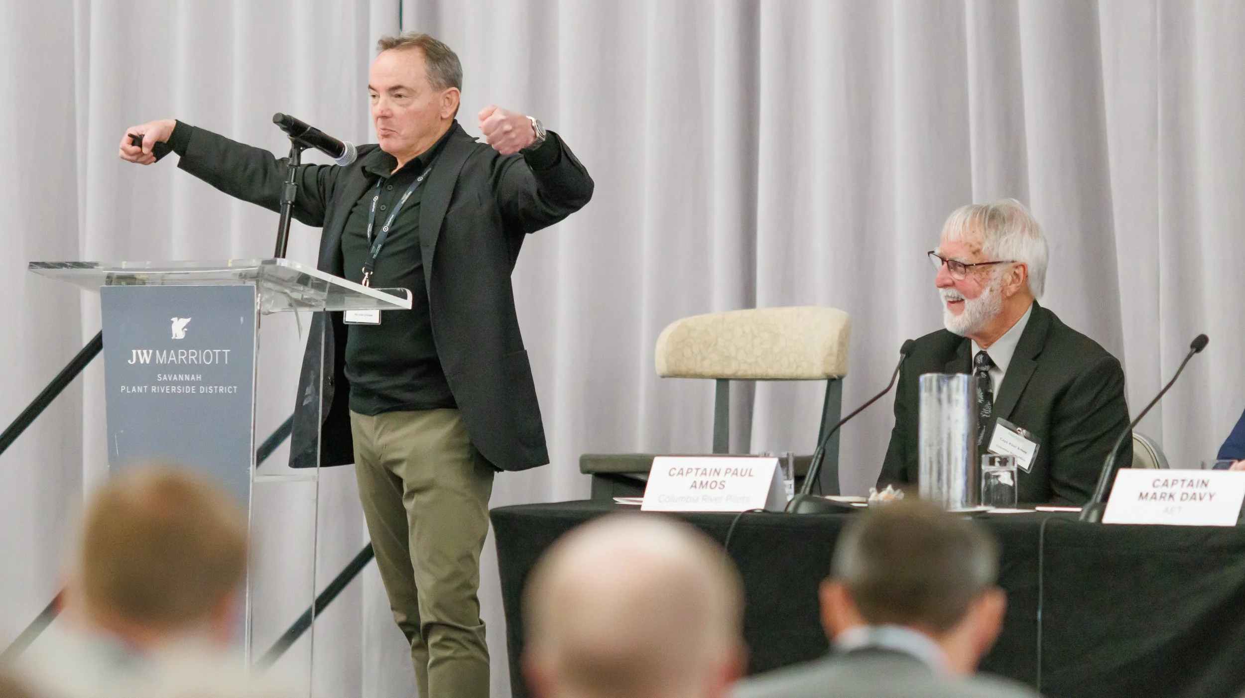 A man at a podium giving a presentation during a conference at JW Marriott in Savannah, with a panel of seated individuals including a man with white hair and glasses, and name tags on the table in front of them.
