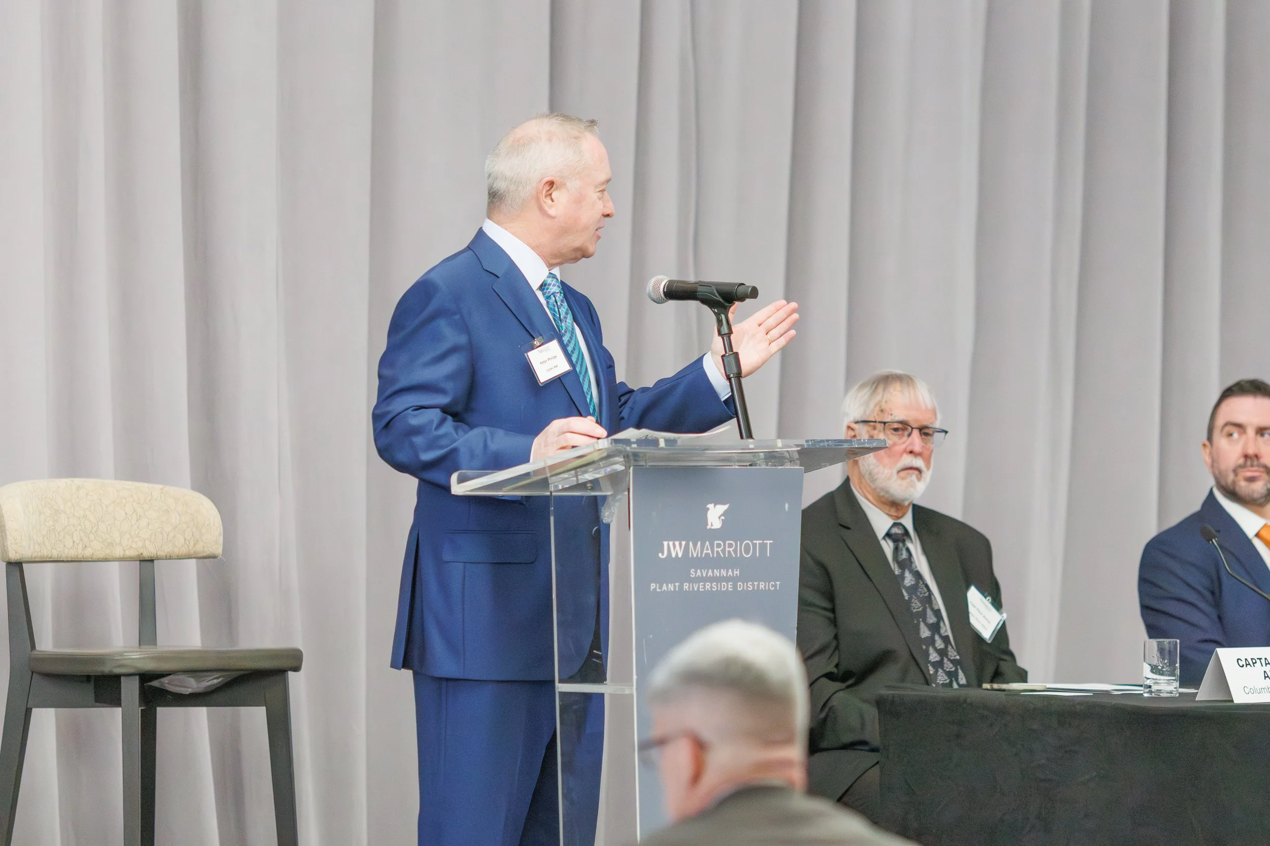 A man in a blue suit and tie is standing at a clear podium, speaking into a microphone at a conference. Behind him, there are two seated men in suits and glasses, one with a beard and the other with gray hair and a beard. The background features ligh