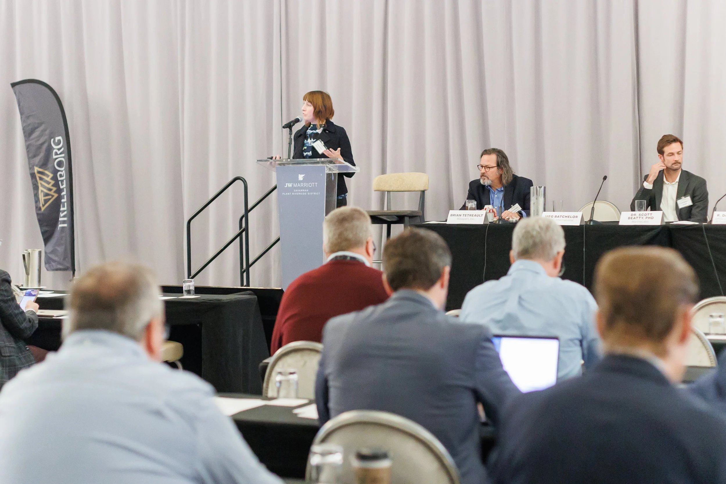 A woman is giving a speech at a conference podium with a JW Marriott sign. Three men are seated at a table behind her, and an audience watches the presentation in a conference room with gray curtains and tables.