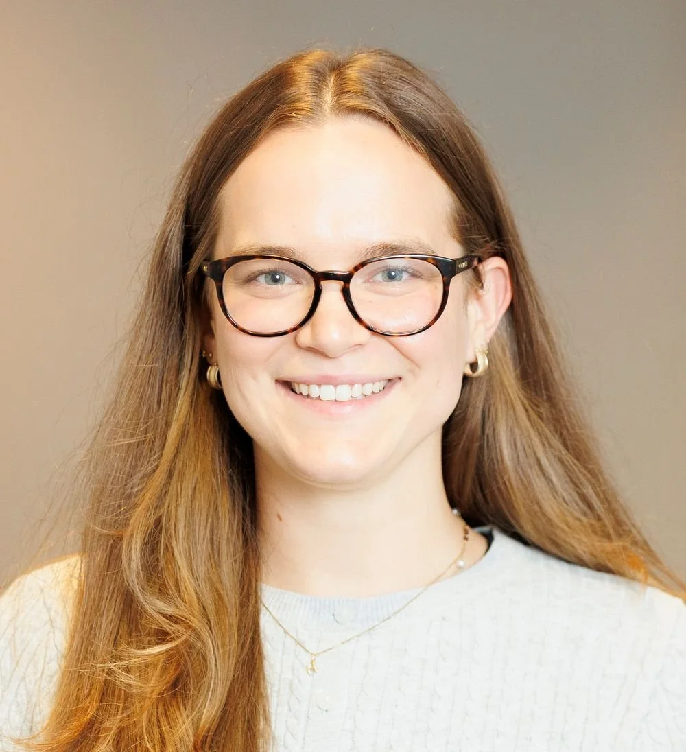 A smiling young woman with long brown hair, wearing glasses, earrings, a necklace, and a light-colored sweater, standing against a plain background.