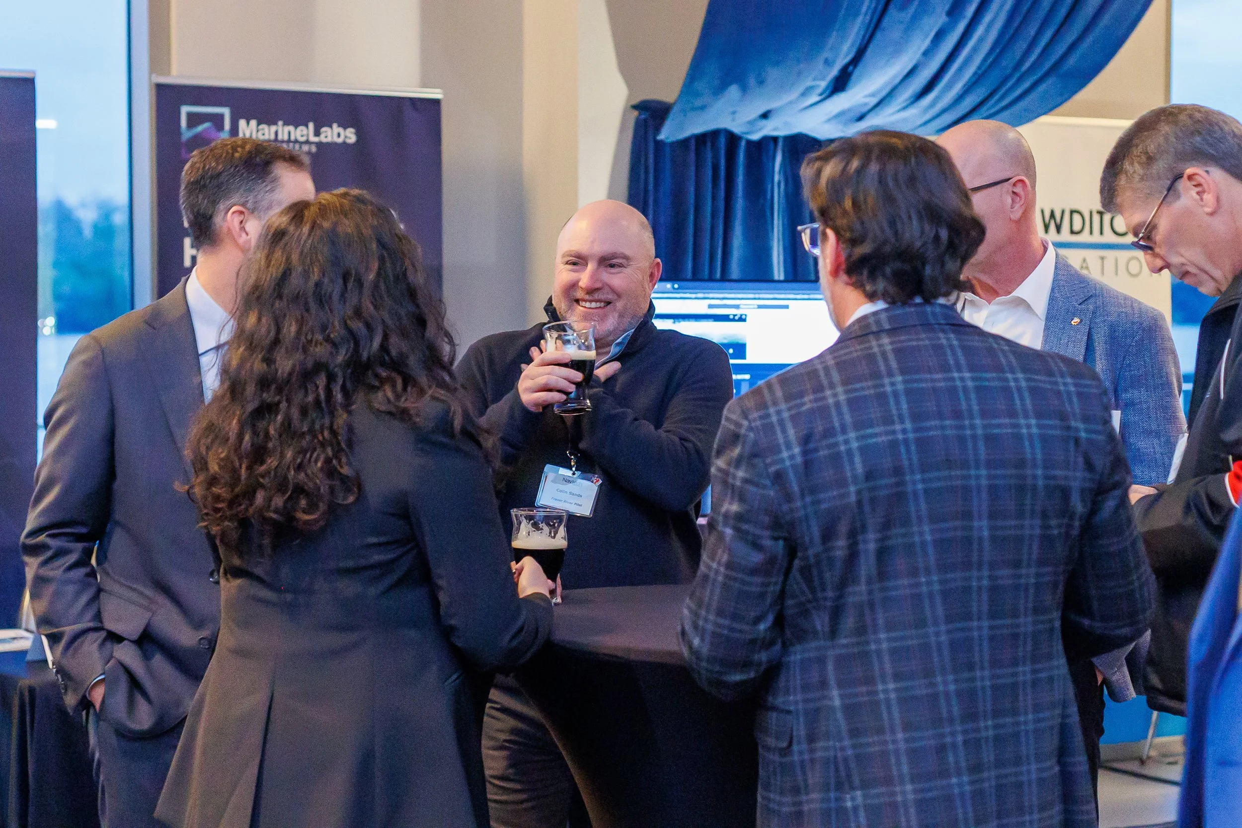 A group of six people in business attire gathered around a high table at a conference or networking event, engaging in conversation with drinks in hand. The background features banners and a monitor.