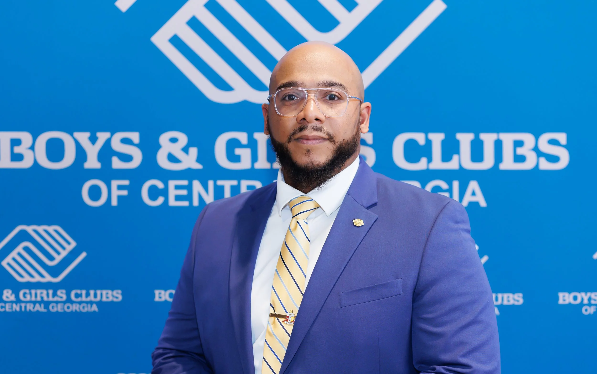 A man in a blue suit and yellow-striped tie stands in front of a blue background with the Boys & Girls Clubs logo and text.