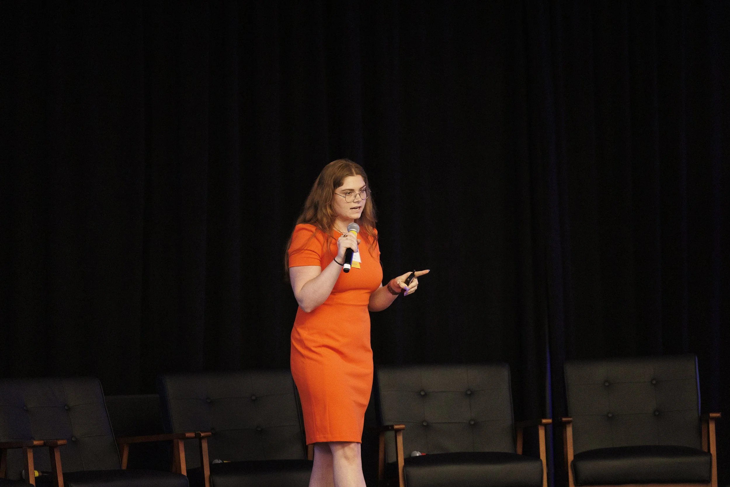 A woman with red hair and glasses wearing an orange dress, speaking into a microphone and gesturing with her left hand on a stage.