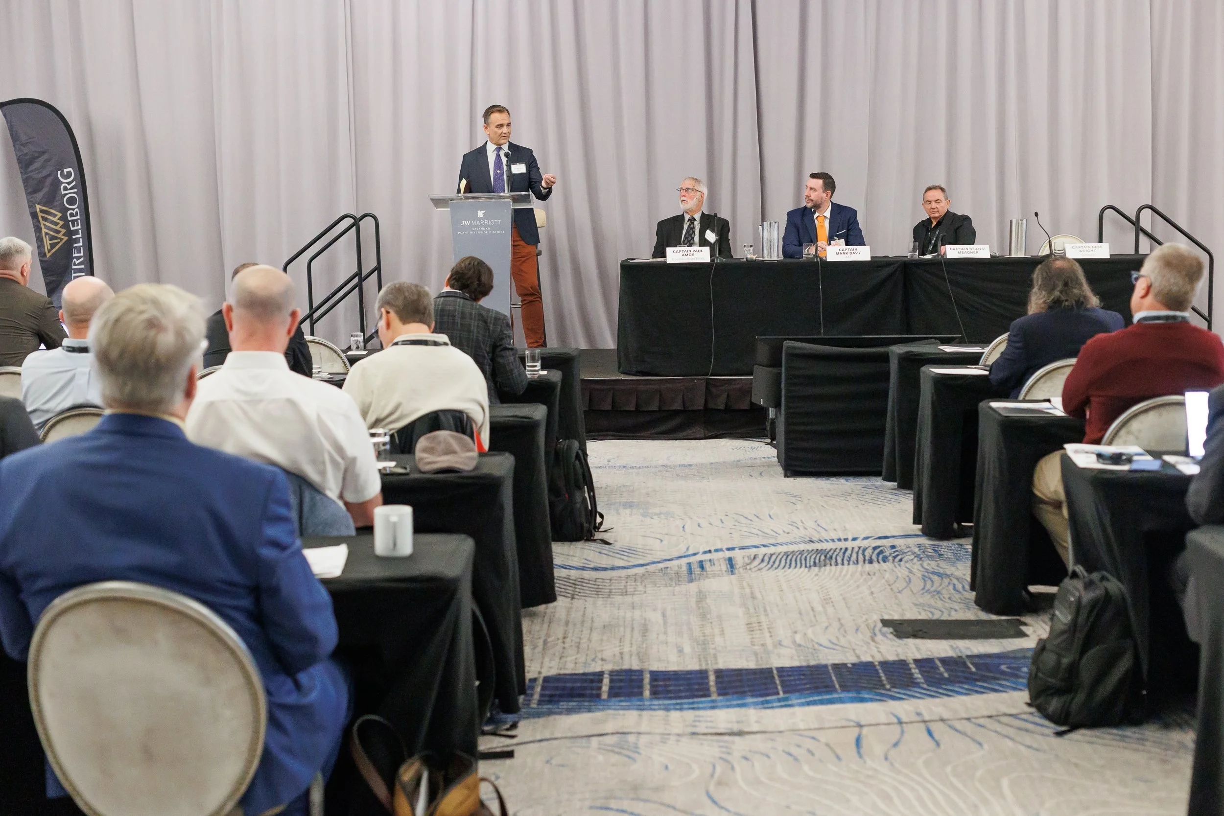 Conference room with a speaker at a podium and panelists sitting at a table, audience members seated at tables facing the stage.